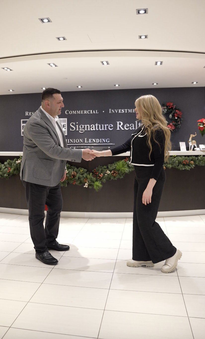 A man and a woman shaking hands in a real estate office decorated for Christmas, with a wreath on the wall and holiday garlands on the reception desk.