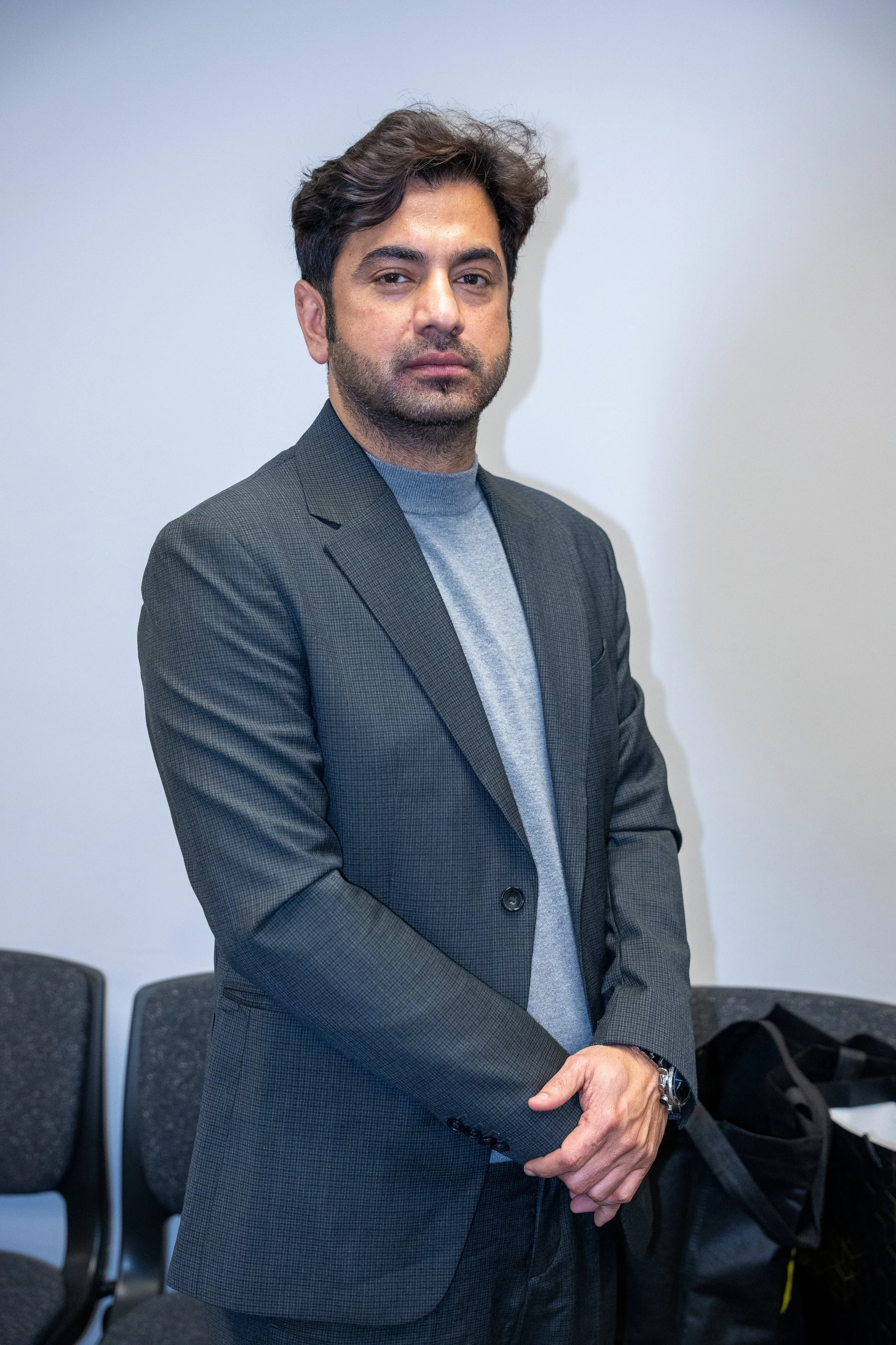 A man with dark hair and a beard wearing a gray blazer and light gray shirt standing in front of a white wall.
