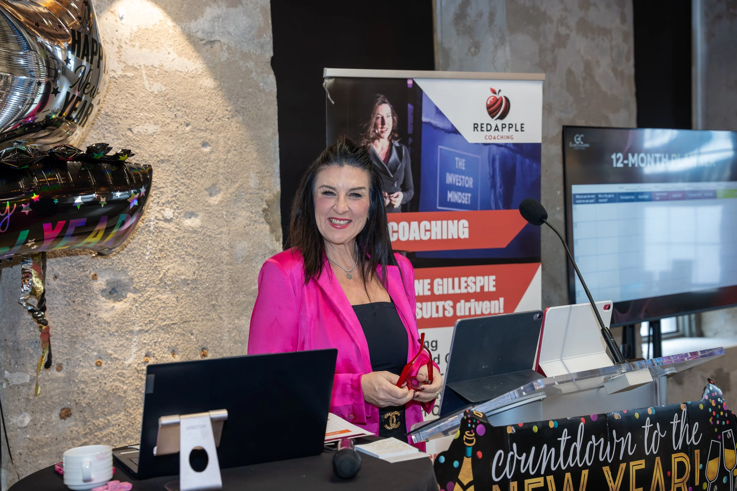 A woman smiling at a New Year's celebration event, standing behind a podium with laptops, balloons, and a banner that says 'Countdown to the New Year'.