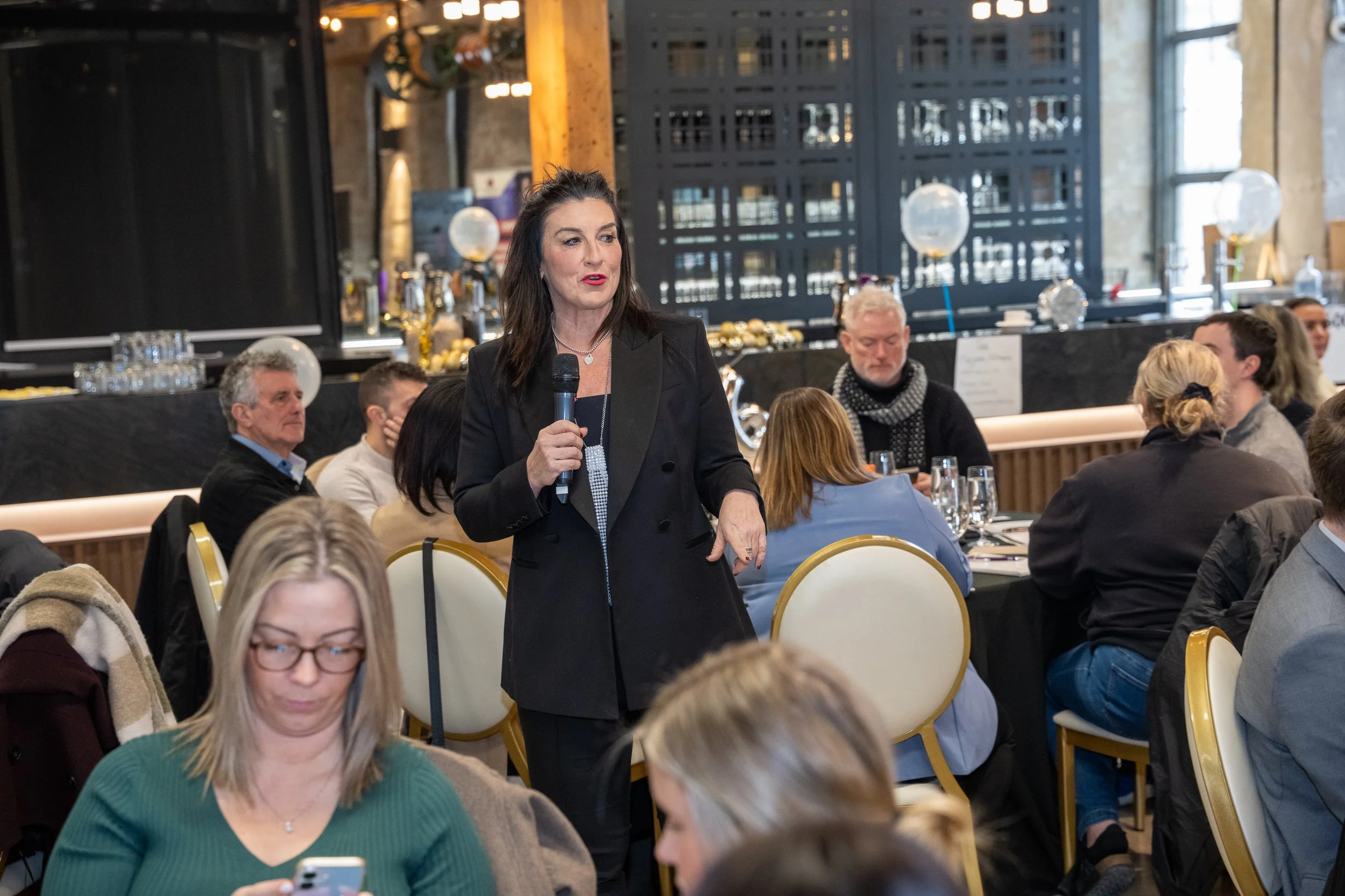 A woman in a black blazer speaking into a microphone at a banquet-style event with attendees seated at round tables, some looking at her and others using their phones, in a well-lit venue with large windows and decorative elements in the background.