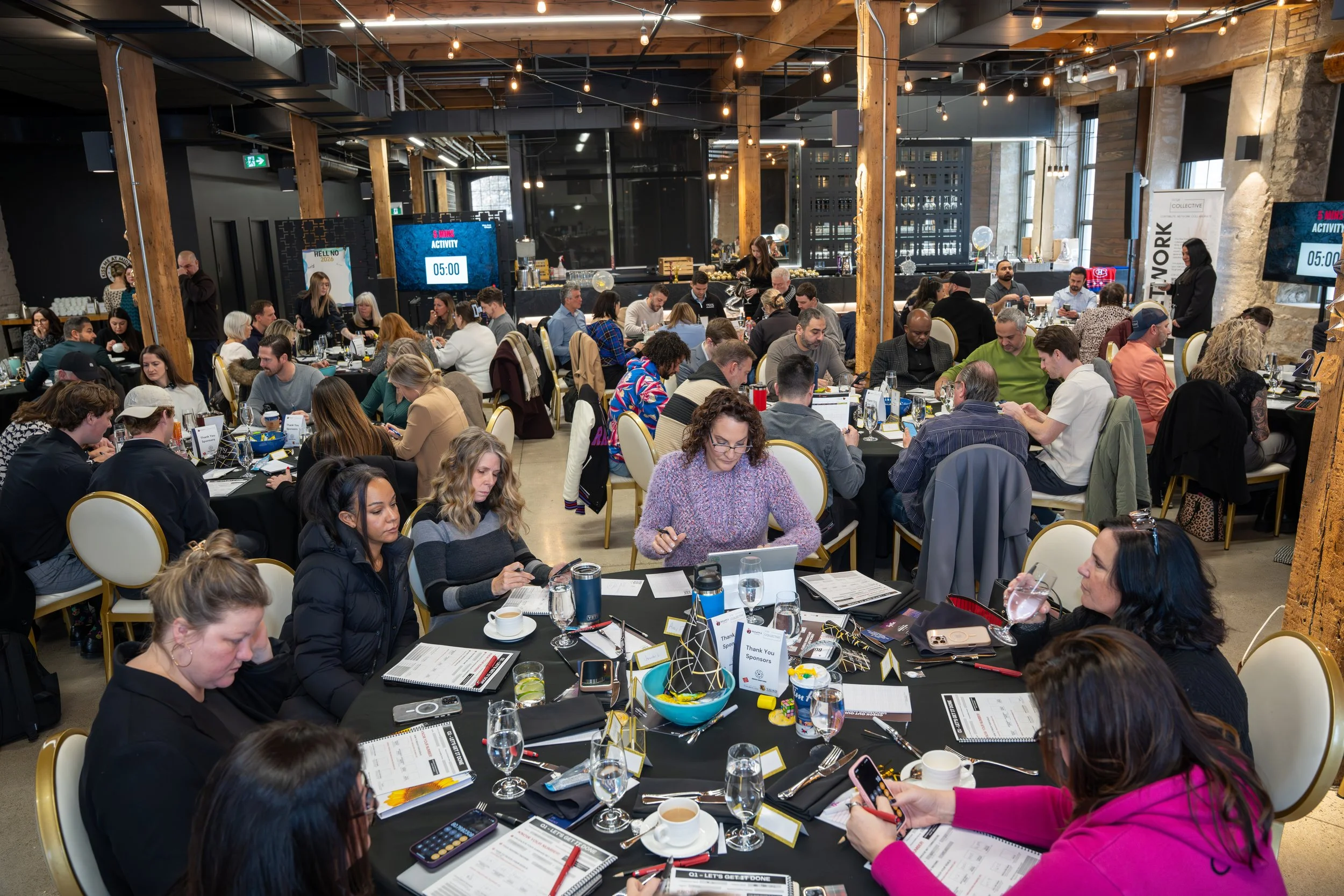 People seated at round tables in a large, well-lit conference or event space with wooden beams, string lights, and large screens displaying a timer and event information.