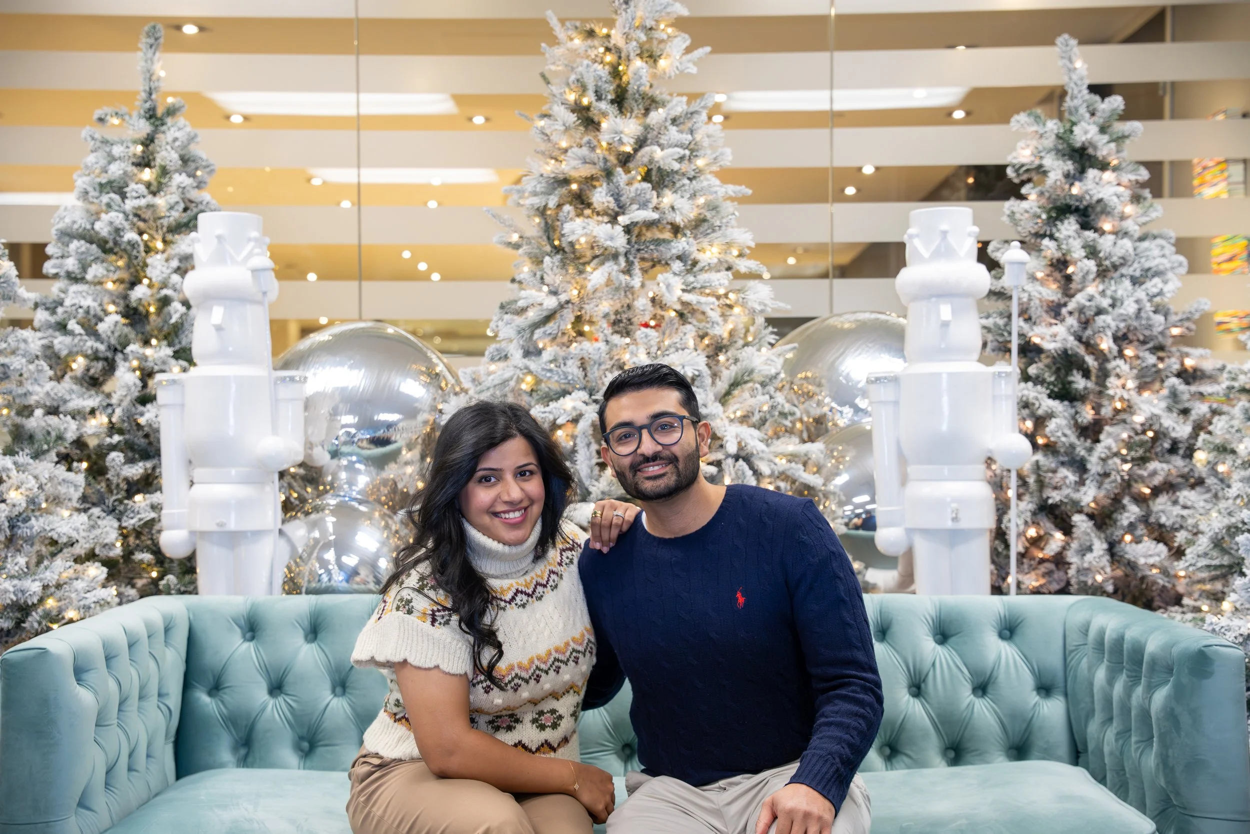 A smiling woman and man sitting on a light blue tufted sofa, posing in front of a decorated Christmas display with snow-covered trees, large silver ornaments, and nutcracker figures.