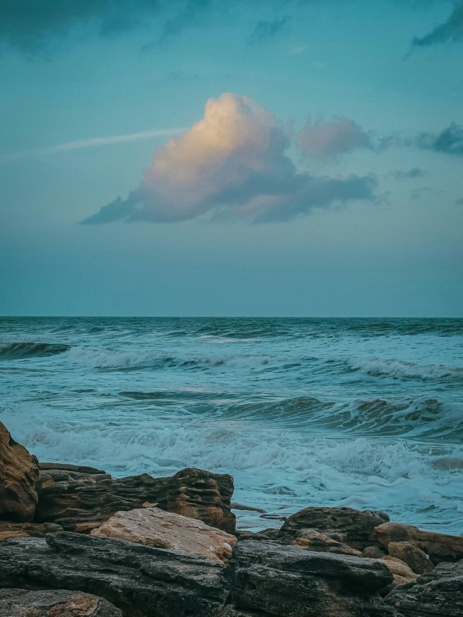 A view of the ocean with waves crashing on rocks in the foreground, a large cloud in the sky, and a blue-green color palette.