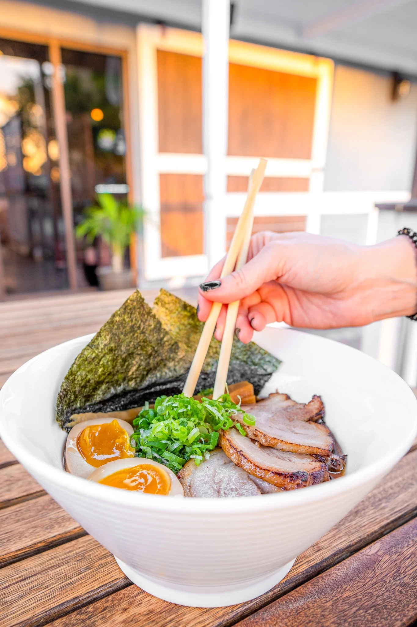 A bowl of ramen with seaweed, soft-boiled eggs, sliced pork, and chopped green onions, with a hand holding chopsticks picking up a piece of pork.