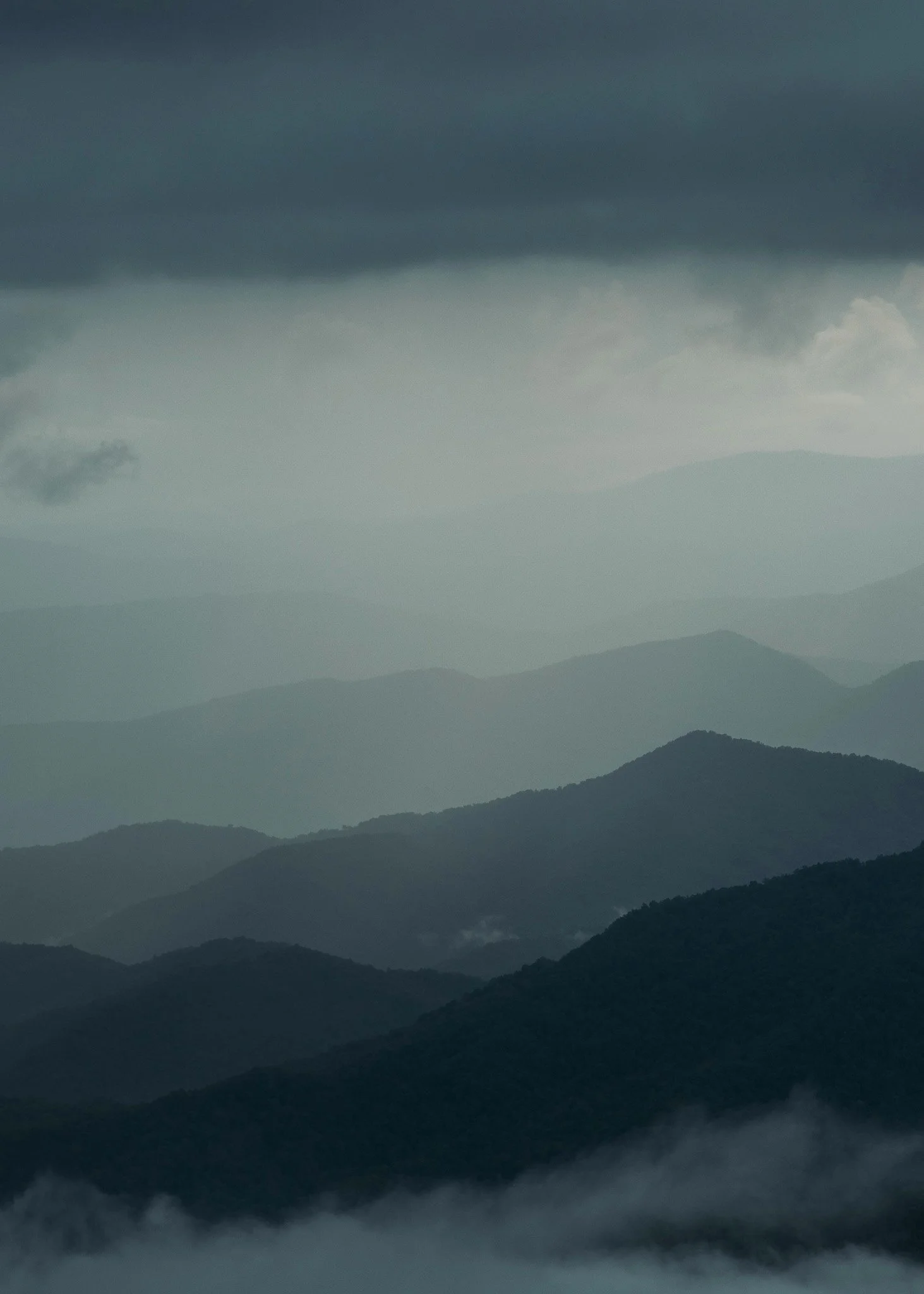 Layered mountain range under dark, cloudy sky with fog at the base of the mountains.