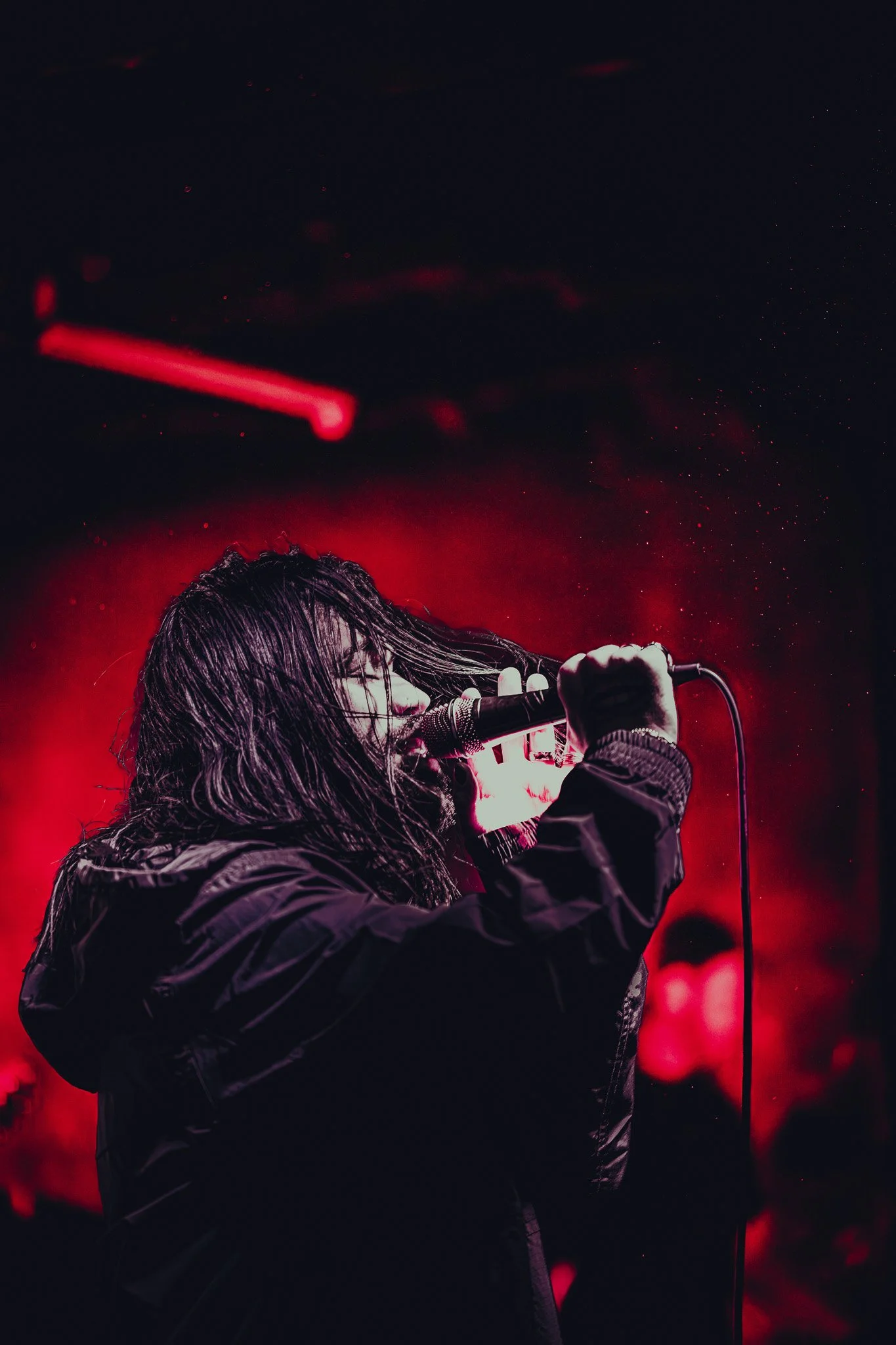A person with long hair singing into a microphone on stage, illuminated by red lighting, with a dark background.