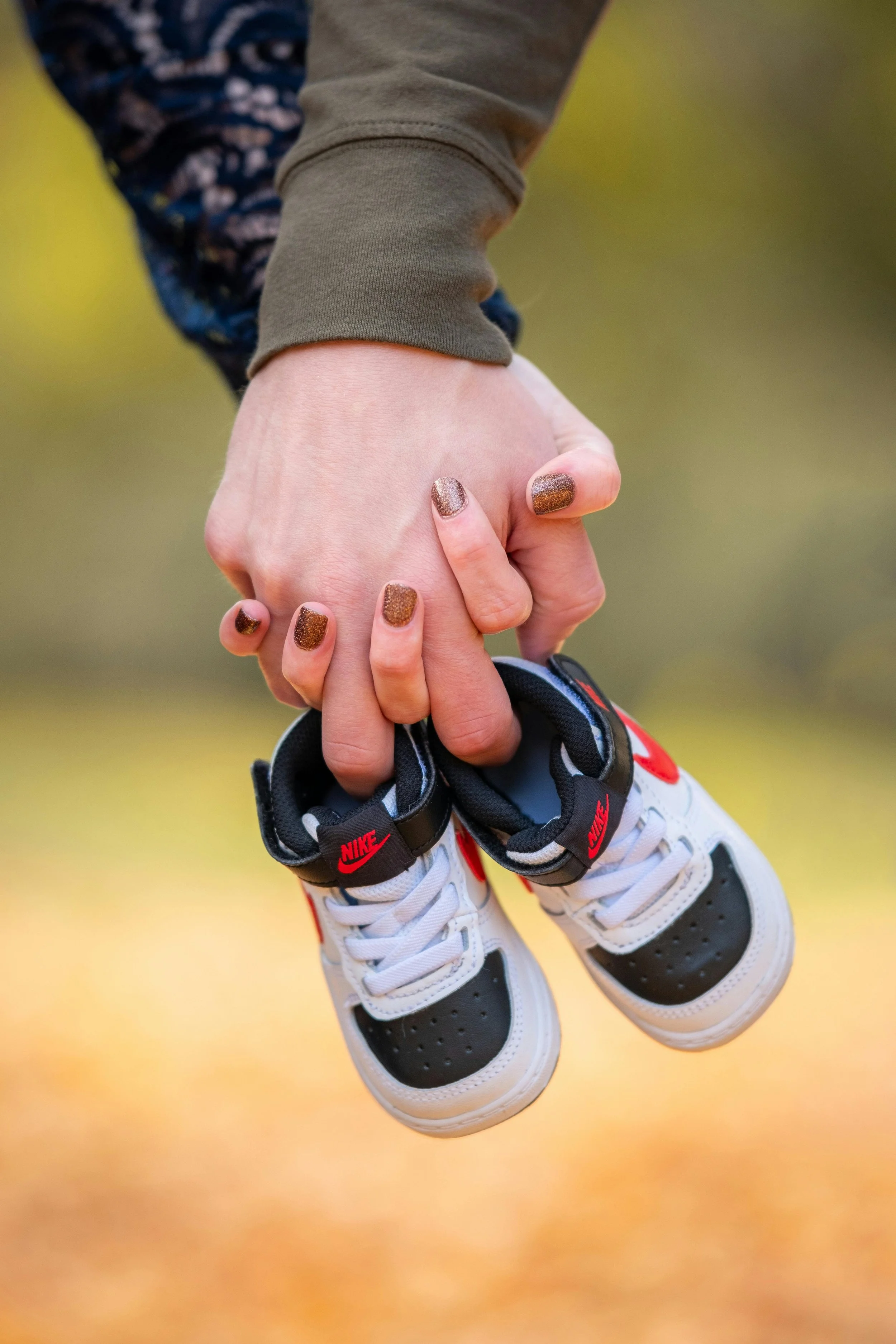 Person holding small Nike sneakers with gold glitter nails, outdoors with blurred yellow and green background.