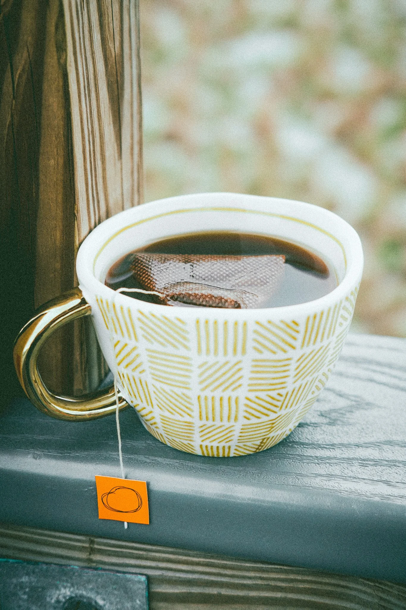 A white coffee mug with a yellow plaid pattern, filled with black coffee and a tea bag inside, sitting on a dark table next to a wooden surface.