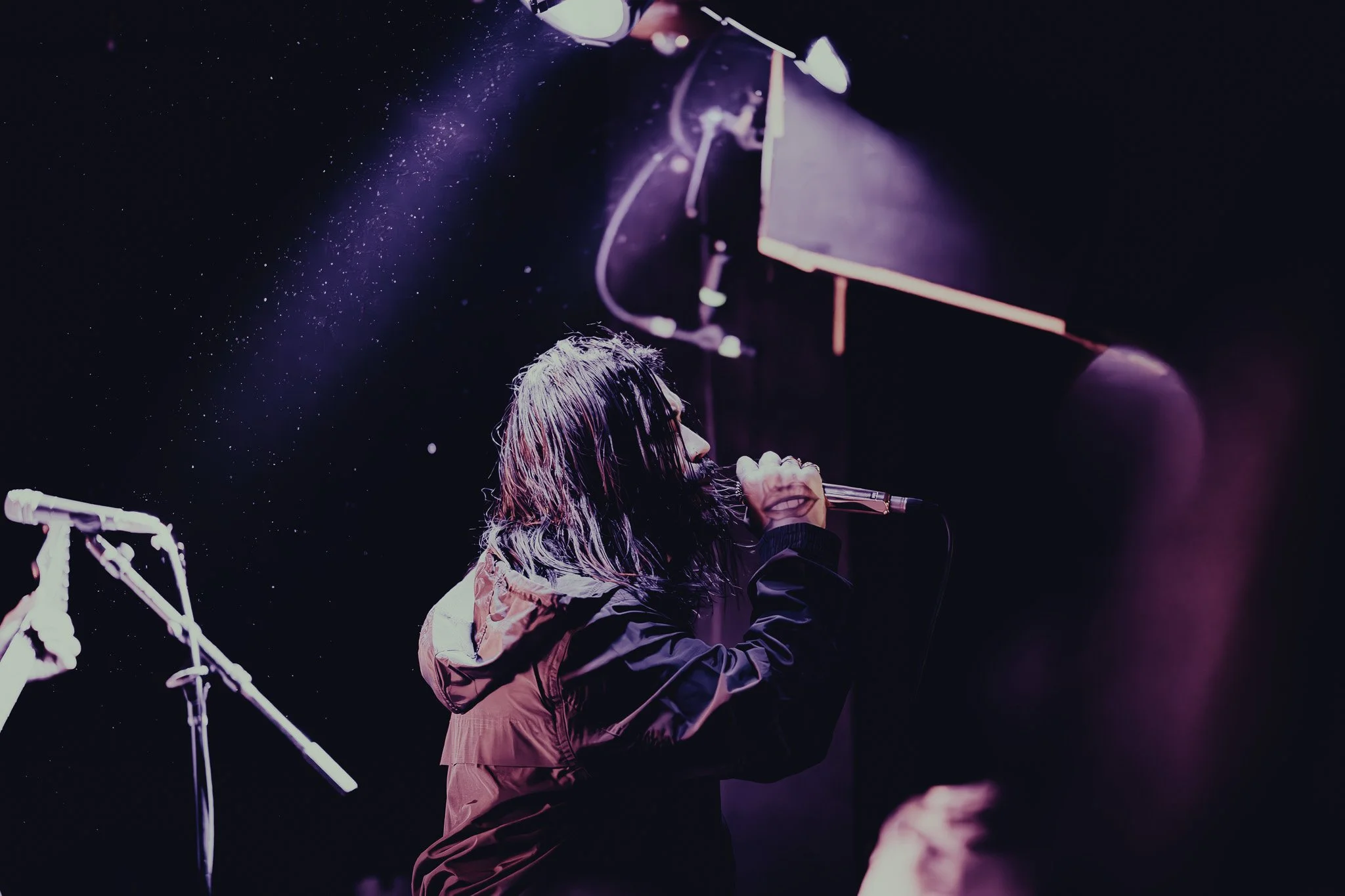 A woman with wet hair singing into a microphone on a dark stage with purple and pink lighting.
