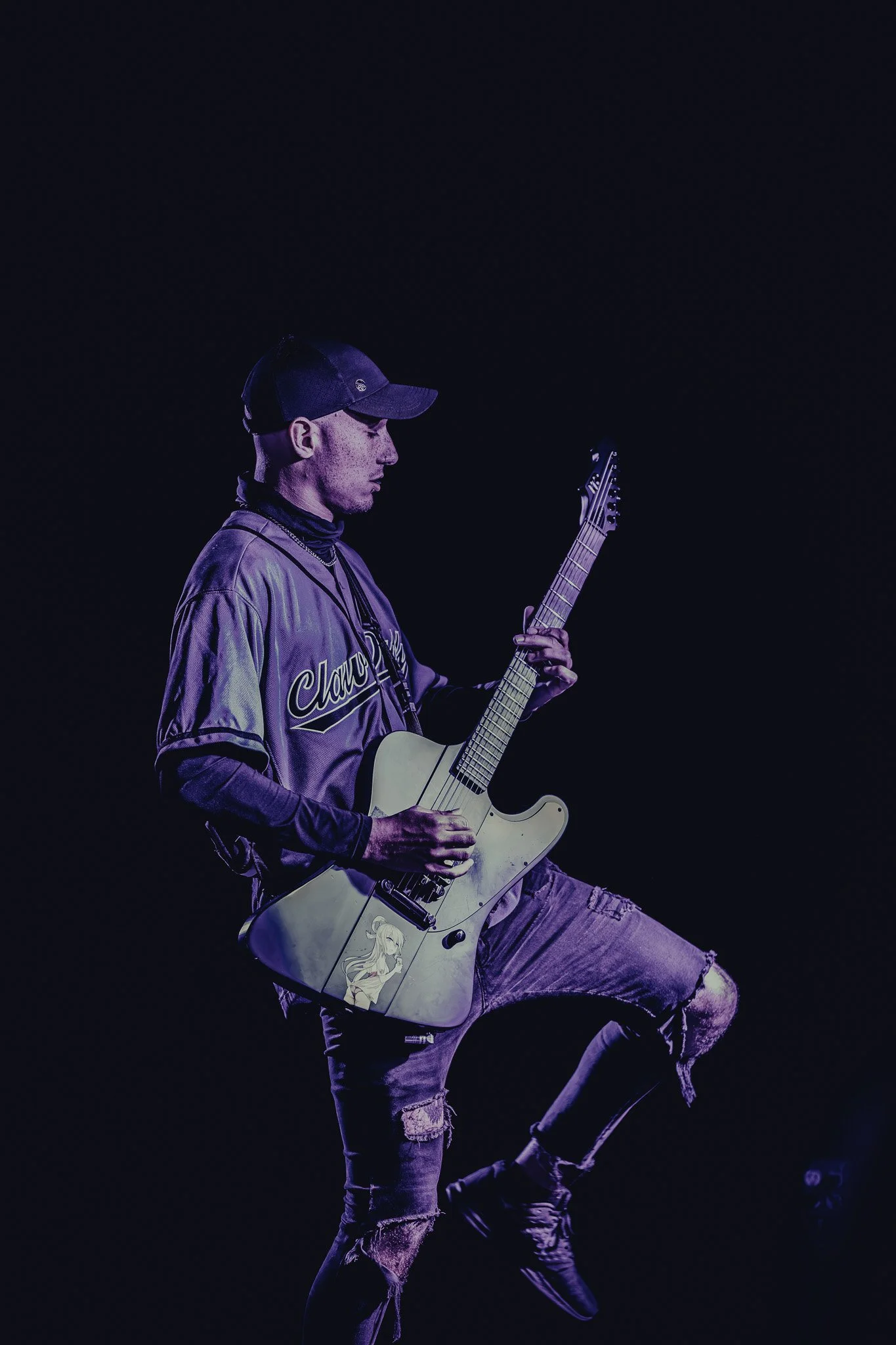 A male guitarist playing an electric guitar on stage with purple lighting, wearing a baseball cap, a jacket, ripped jeans, and sneakers.
