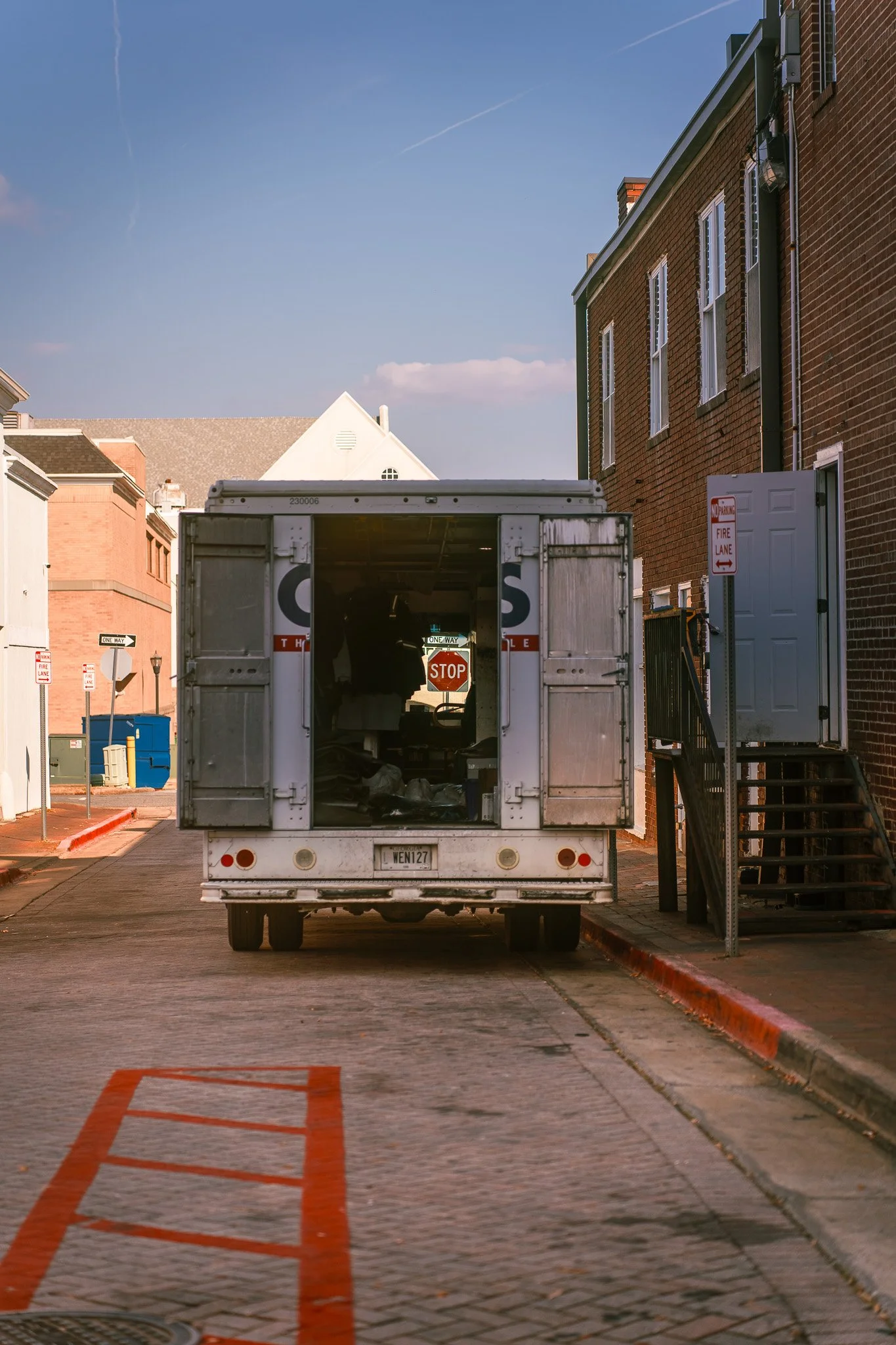 A delivery truck parked on a cobblestone street with a staircase leading to an entrance on the right, surrounded by brick and painted buildings, under a blue sky with some clouds.