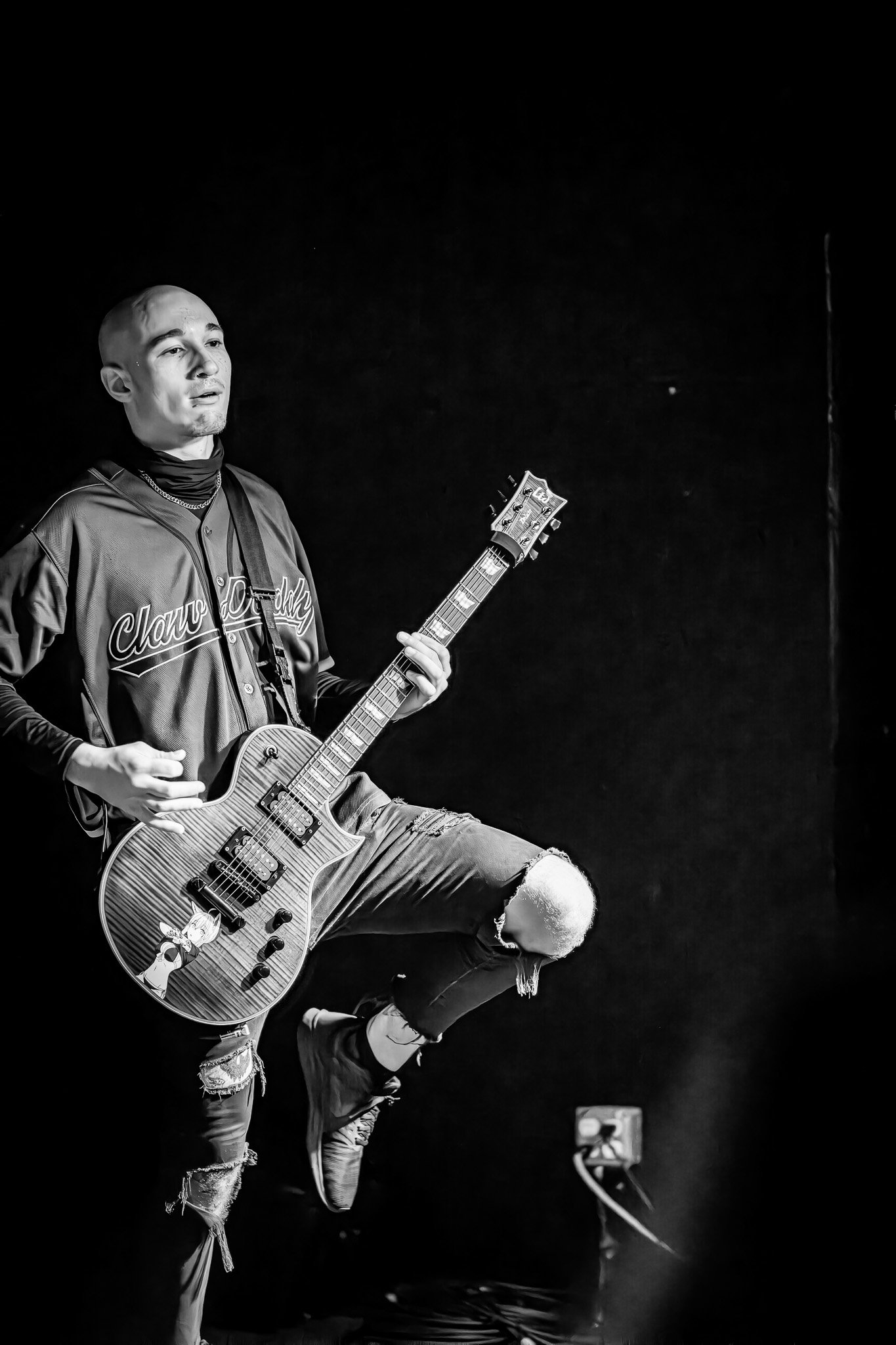 A young man playing an electric guitar on stage, dressed in a sports jersey and ripped jeans, with a black background.