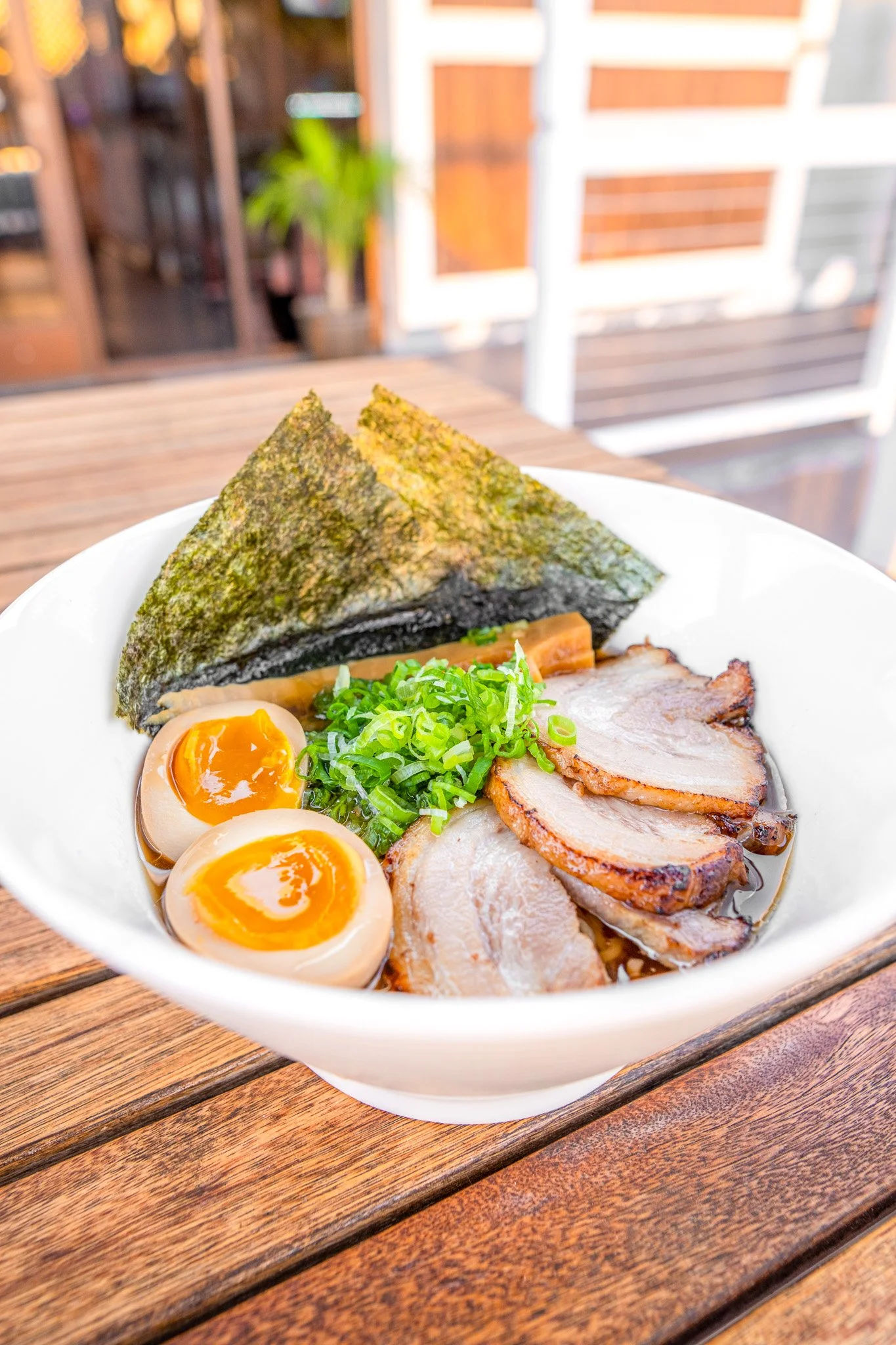 A bowl of ramen with seaweed, soft-boiled eggs, sliced pork, and chopped green onions placed on a wooden table outdoors.