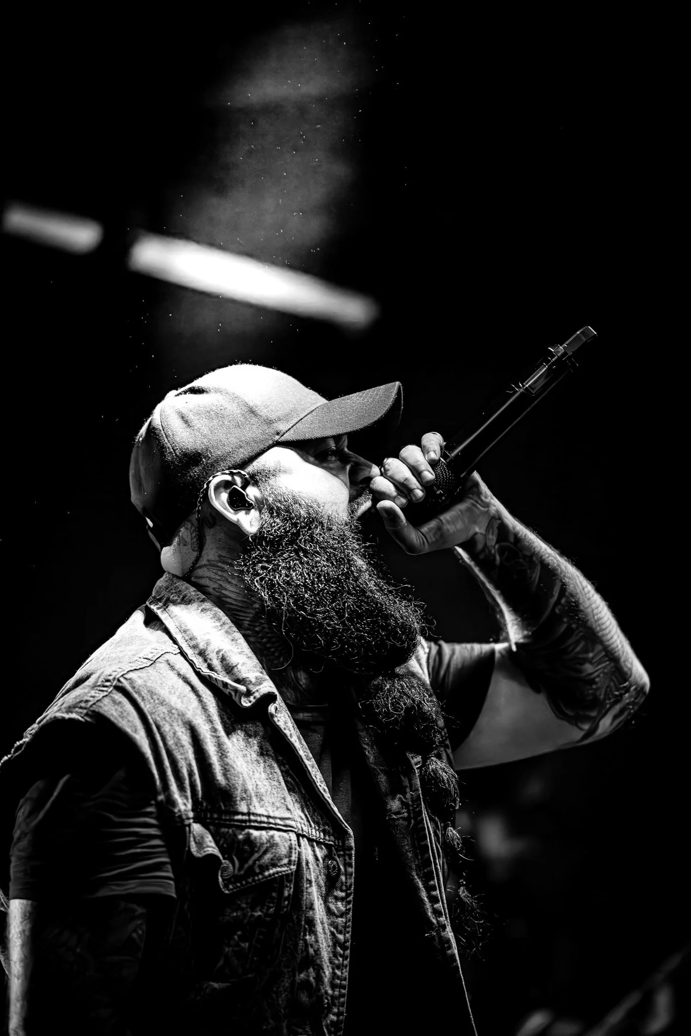 A black and white photo of a male singer performing on stage, wearing a baseball cap, with a beard, holding a microphone close to his mouth, and wearing headphones. He is dressed in a sleeveless denim vest and has tattoos on his arms.