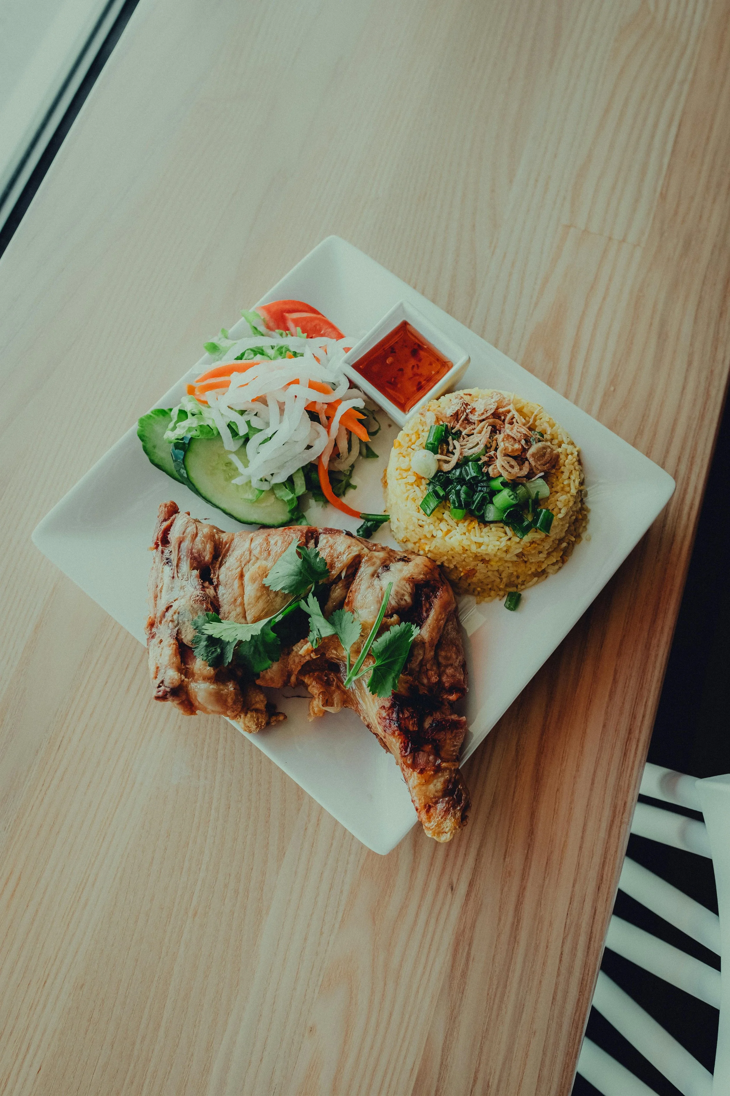 Plate with grilled chicken, Vietnamese-style fried rice, and fresh salad with vegetables, accompanied by dipping sauce, on a wooden table.