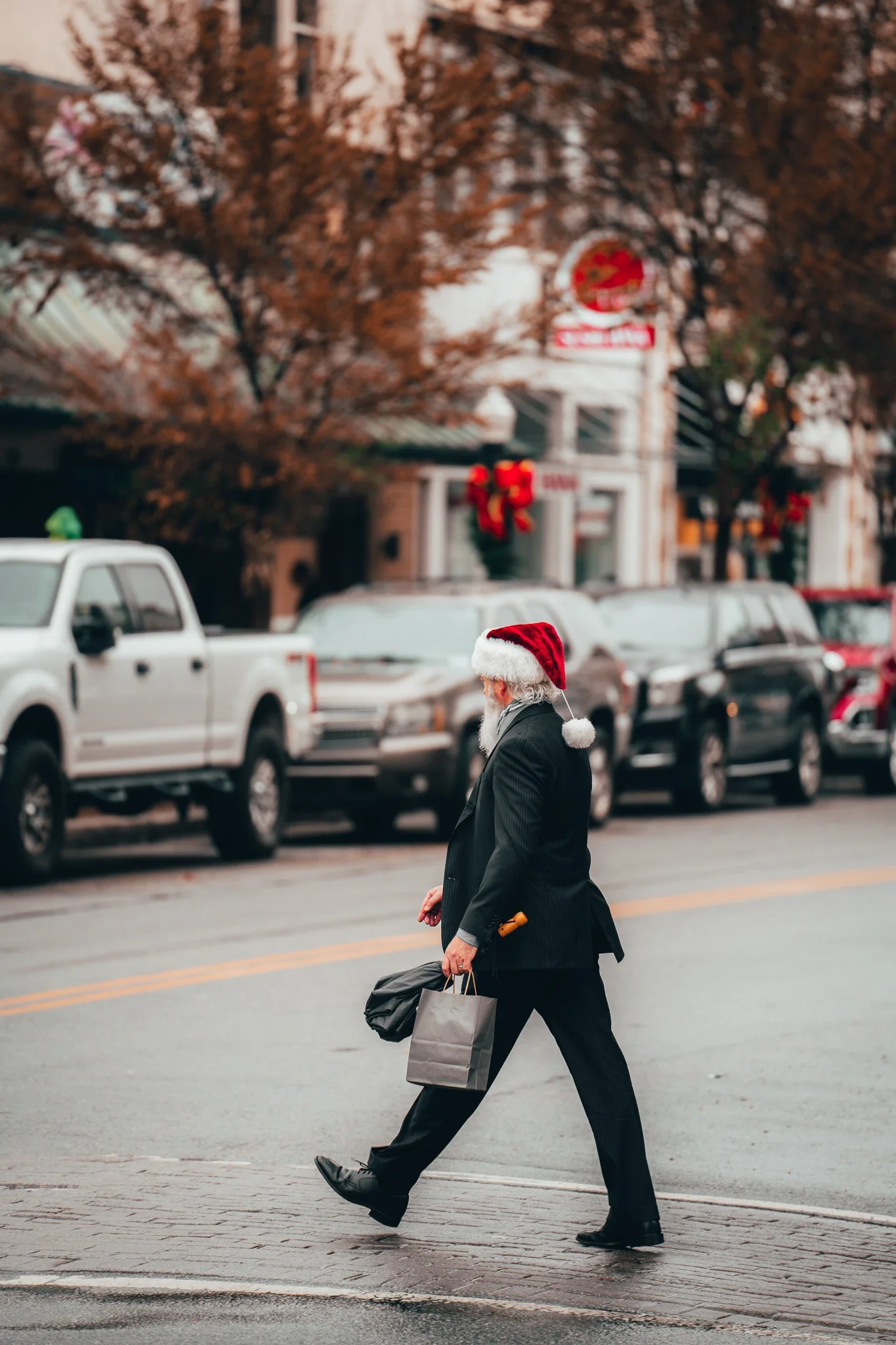 A man dressed in a business suit and Santa hat walks across a city street carrying shopping bags and a small wrapped gift, with parked cars and holiday decorations in the background.