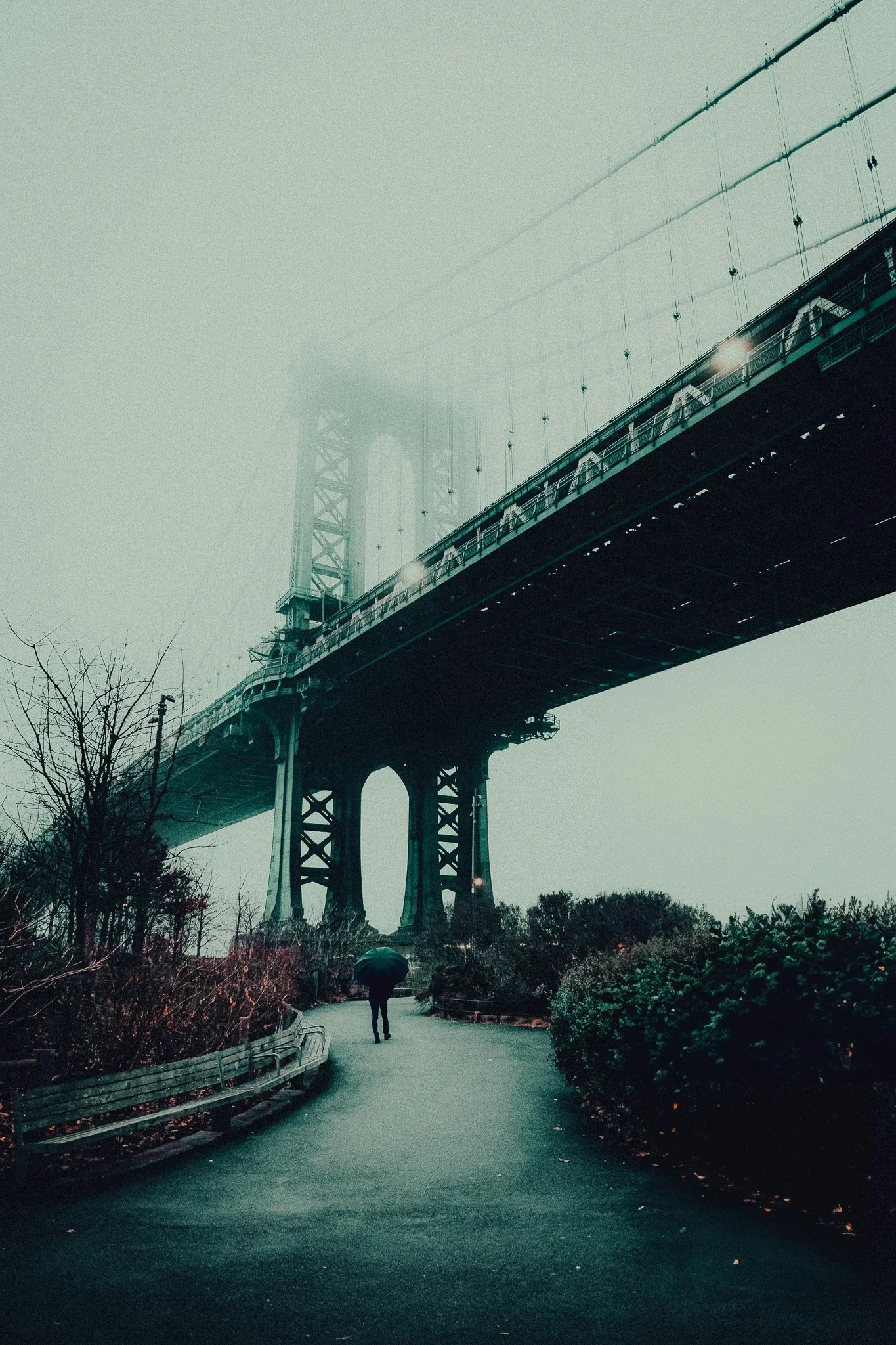 A person with an umbrella walking along a pathway under a bridge on a foggy day.