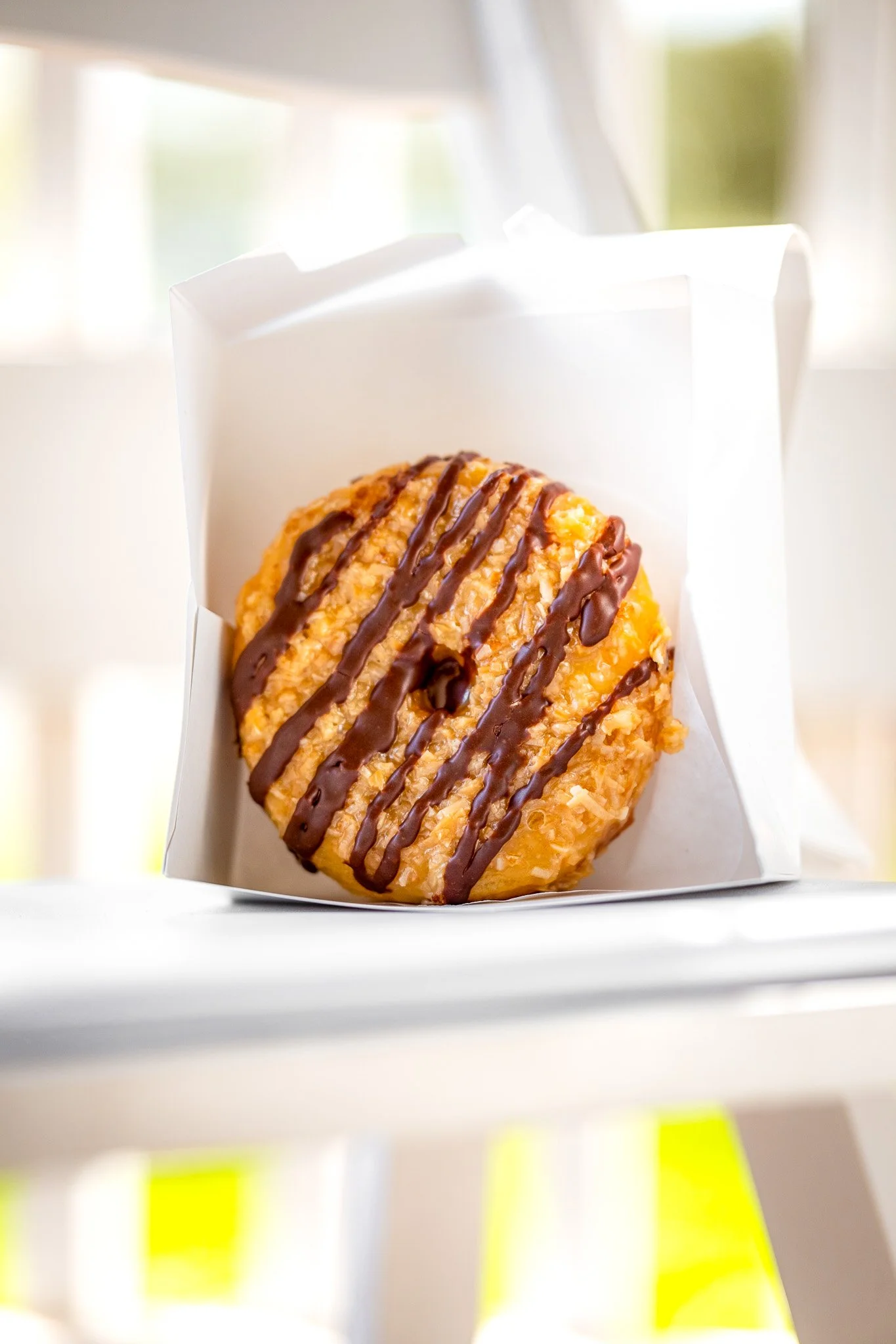 A round pastry drizzled with chocolate, placed inside a white paper box, with a blurred background of an indoor setting with windows.