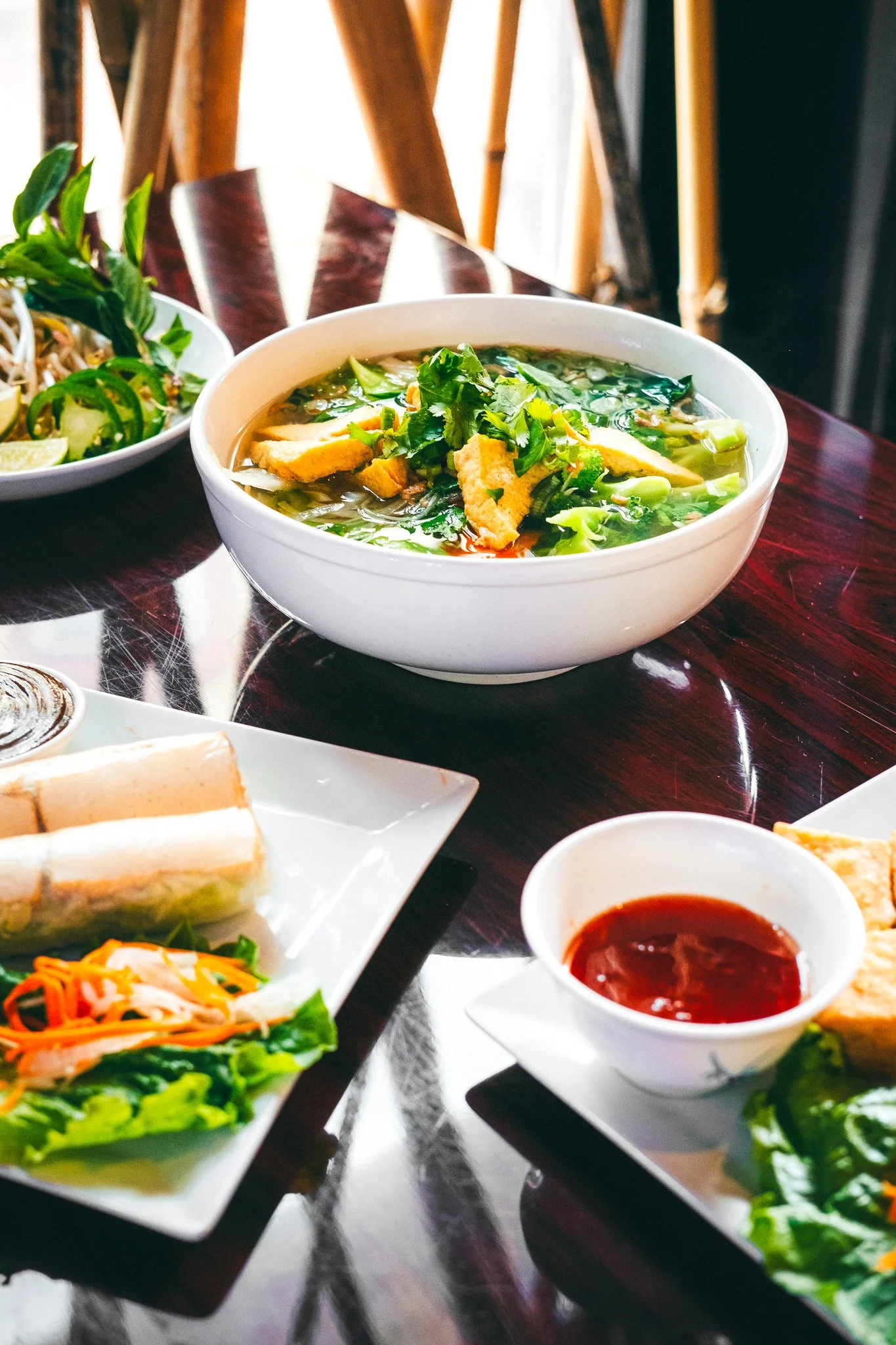 A bowl of Vietnamese pho with herbs and green onions, surrounded by salads, spring rolls, and a small bowl of red dipping sauce on a dark wooden table.