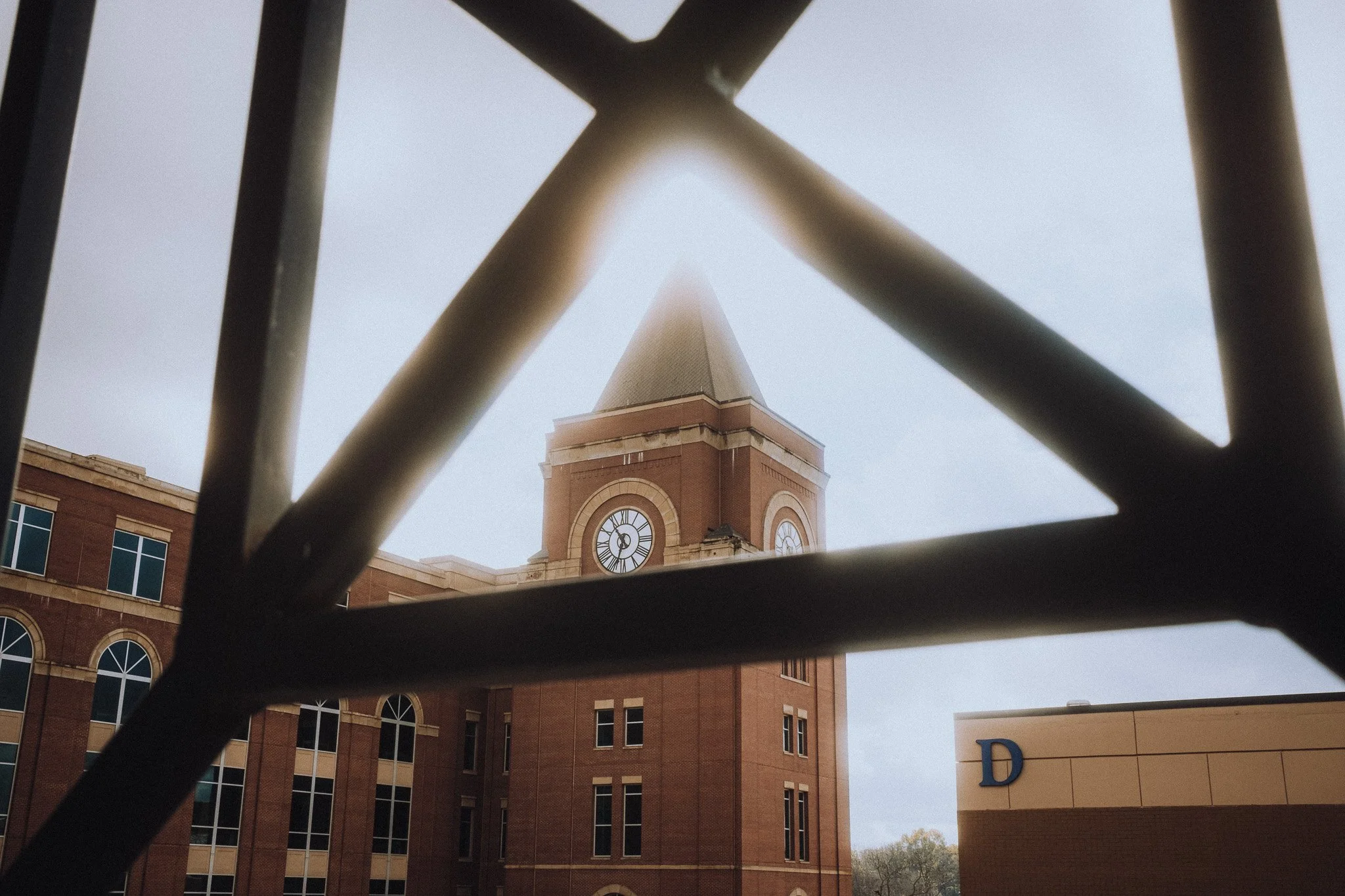 A clock tower seen through a metal grid, with part of a building marked with a letter D on the right.