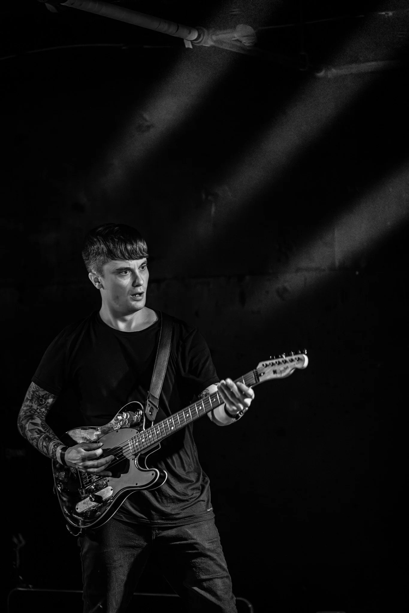 A young man playing an electric guitar on stage, illuminated by stage lights in black and white.