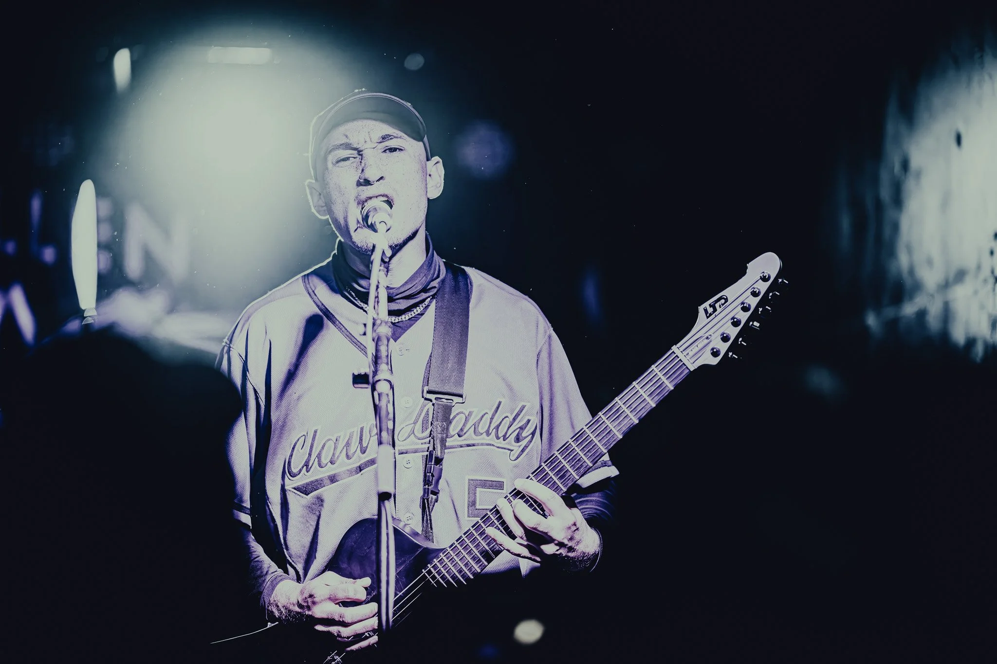 Male musician singing into a microphone while playing an electric guitar, wearing a baseball cap and a sports jersey, with stage lighting and a dark background.