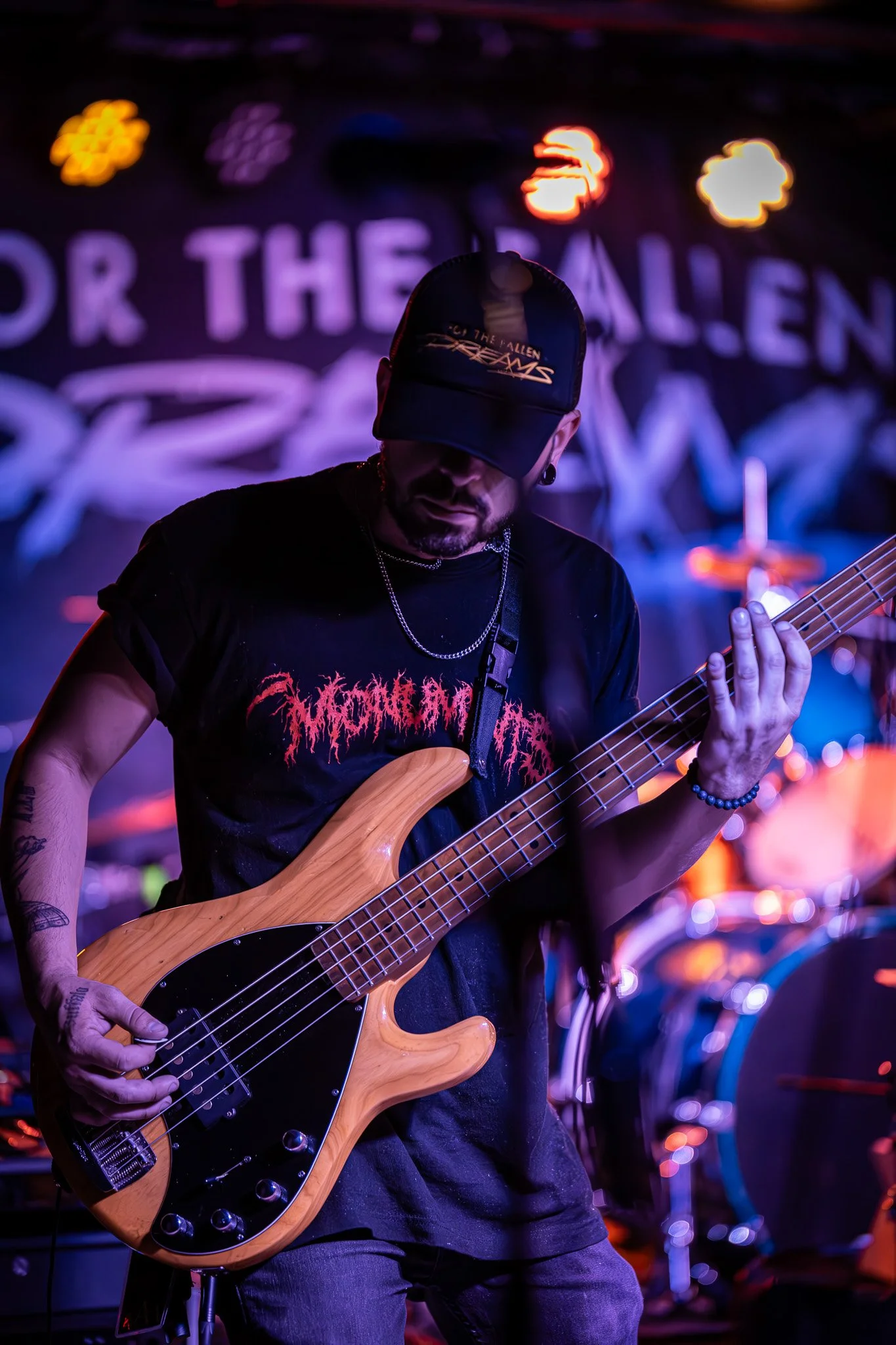 A musician playing an electric bass guitar during a live performance, wearing a black cap and T-shirt with red text, with colorful stage lights and drums in the background.