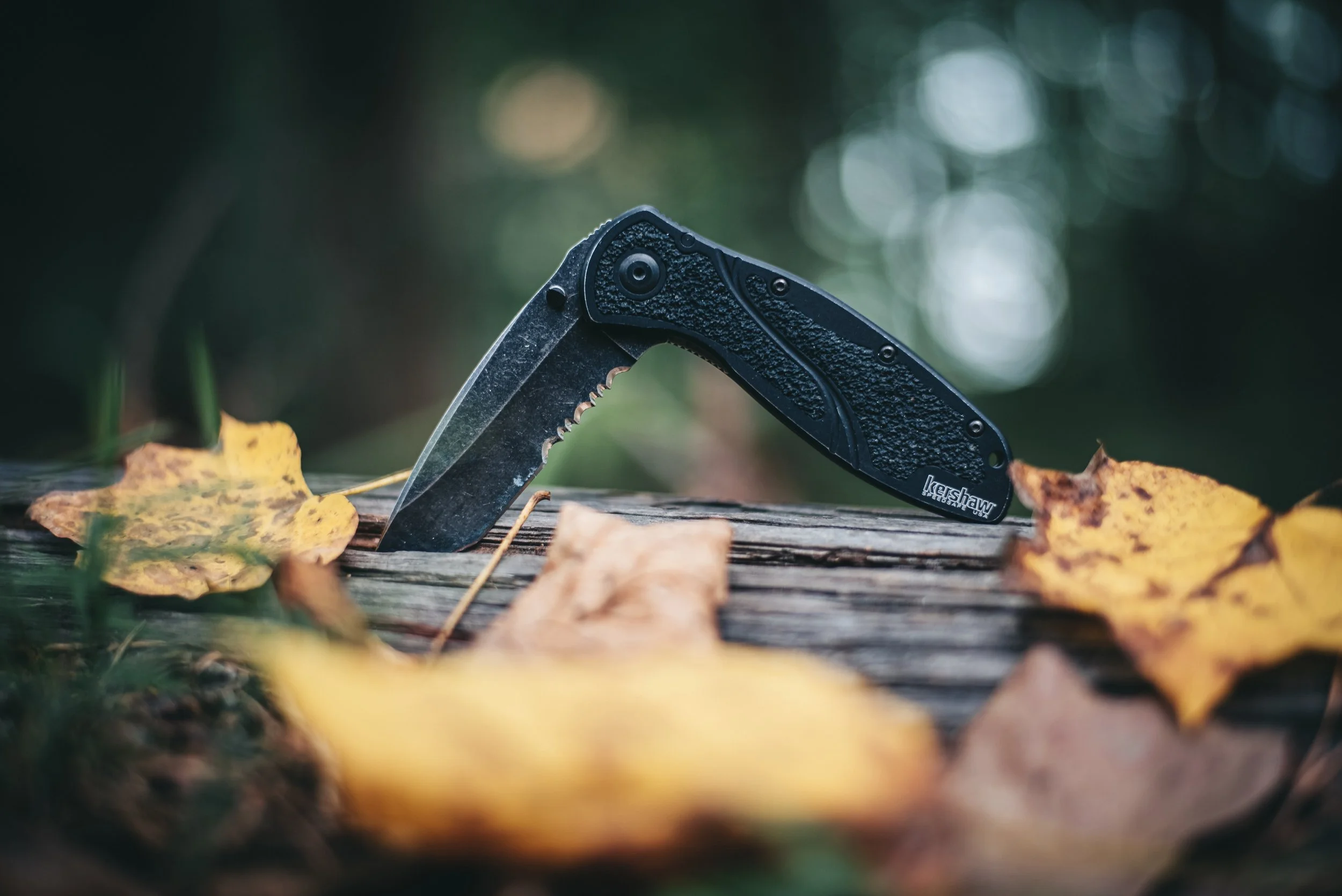 A black multi-tool with a serrated blade is partially opened and resting on a weathered wooden surface, surrounded by yellow autumn leaves in an outdoor setting.