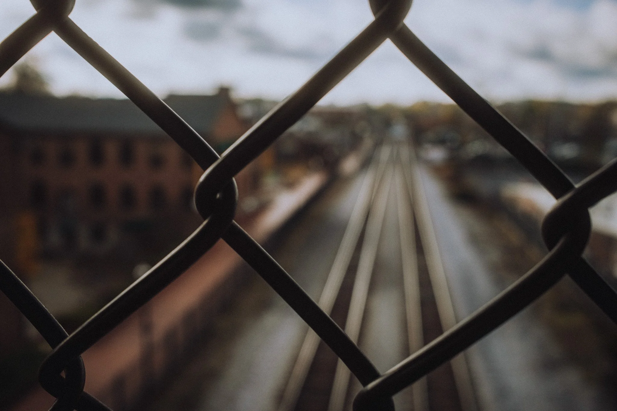 View of railway tracks through a chain-link fence with blurred buildings and sky in the background.