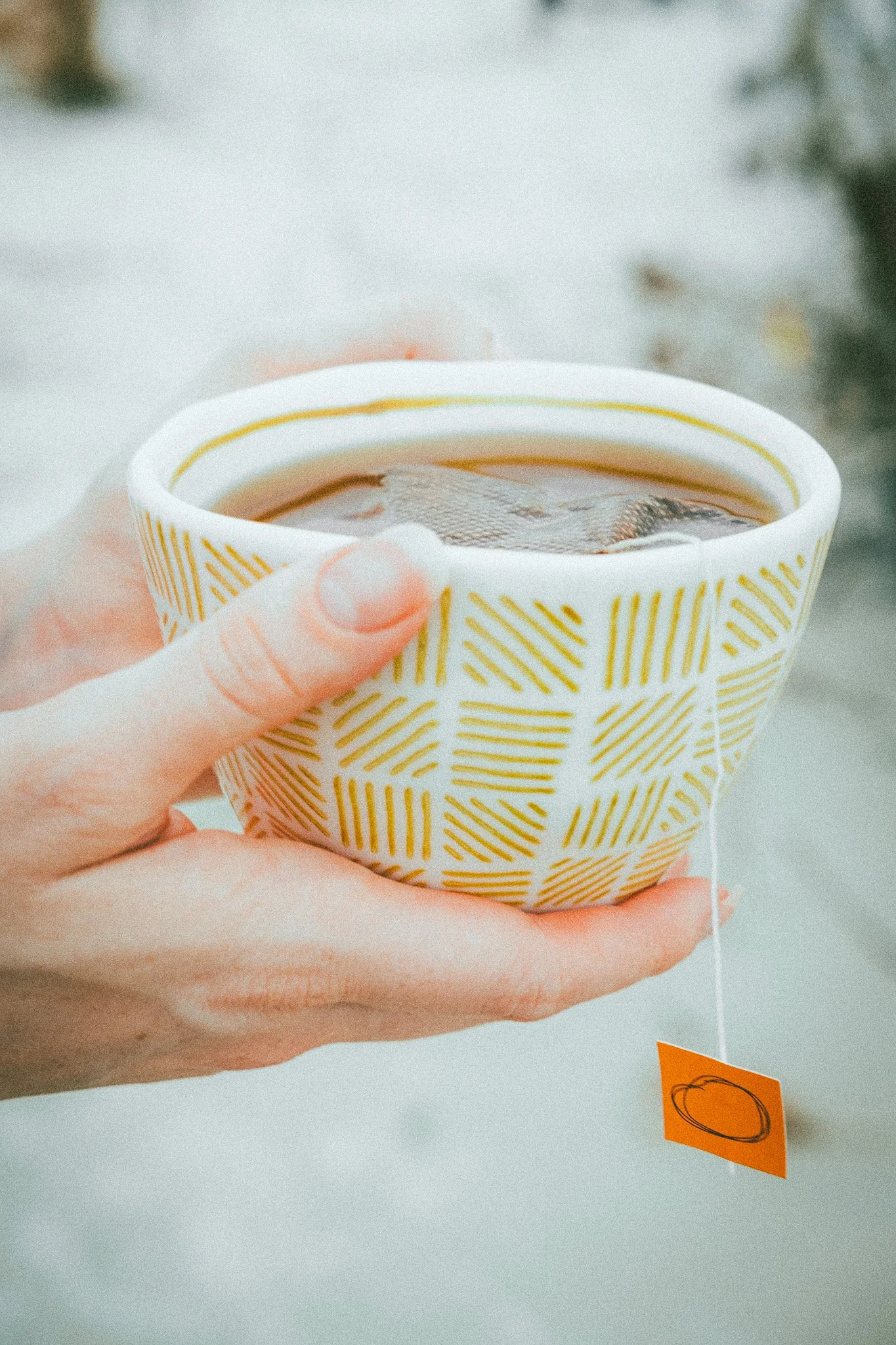 A hand holding a ceramic cup with a yellow geometric pattern, filled with hot tea and a tea bag tag hanging outside.