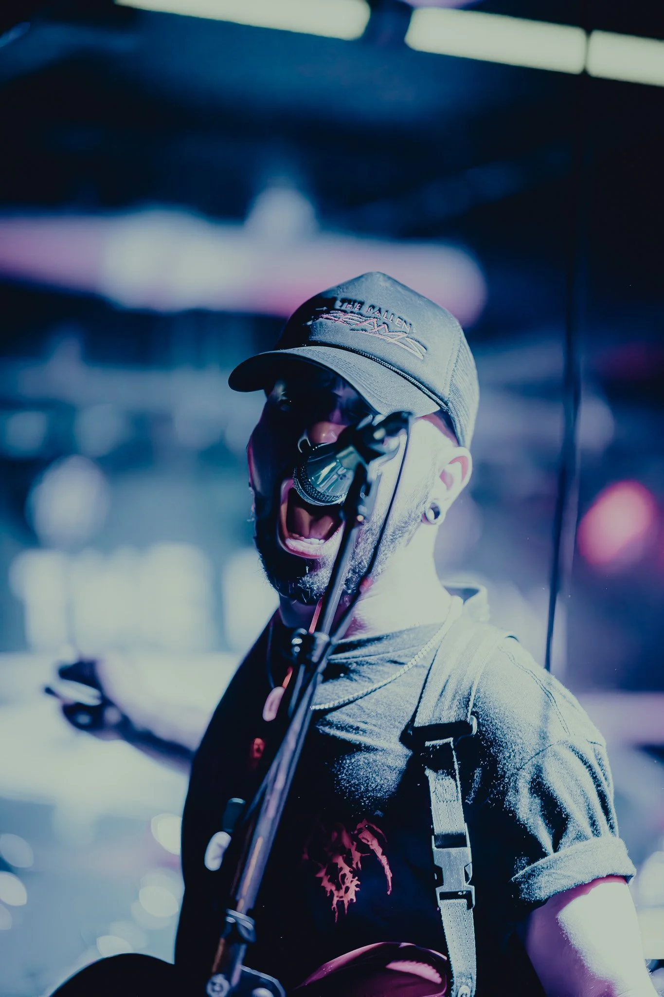 A man in a baseball cap and sunglasses singing passionately into a microphone on stage, with bright stage lights and blurred background.