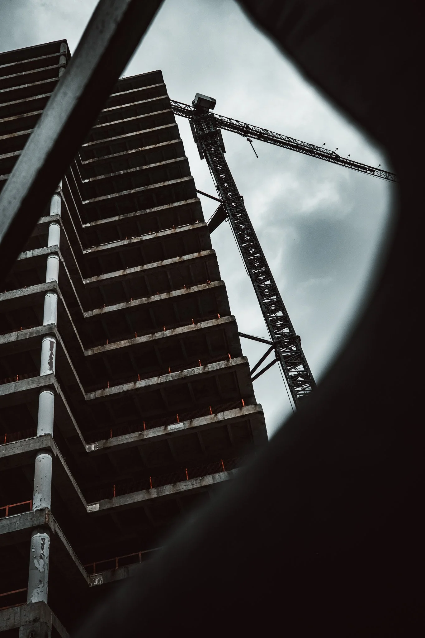 Looking up at a construction site with a tall building under construction and a crane against a cloudy sky, framed by dark metal structures in the foreground.