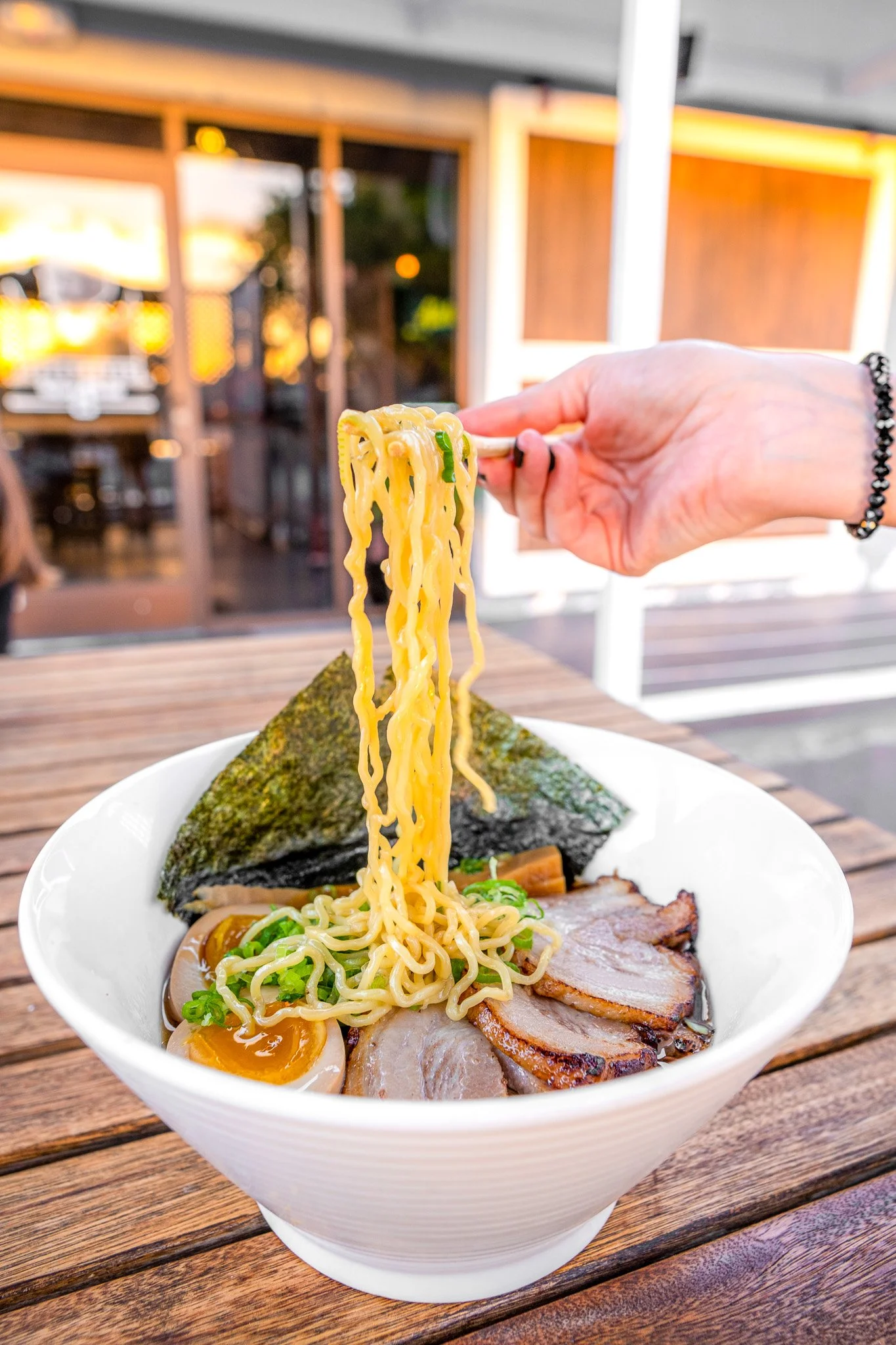 A bowl of ramen with sliced pork, a soft-boiled egg, seaweed, green onions, and noodles being lifted with chopsticks.