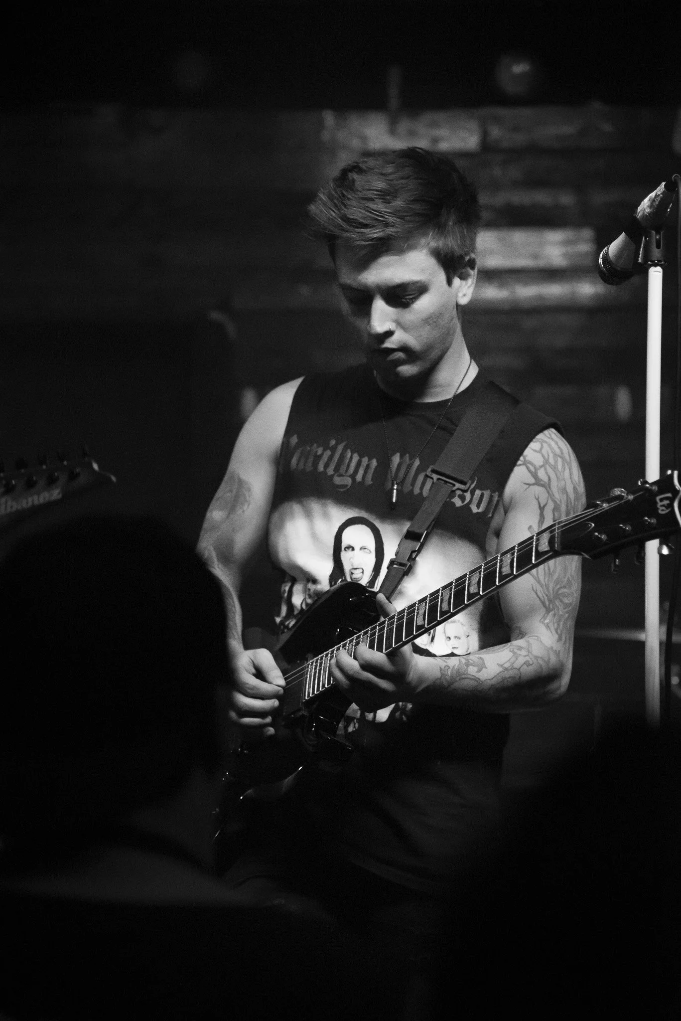 A young man with tattoos on his arms playing an electric guitar during a live performance in a dimly lit venue.