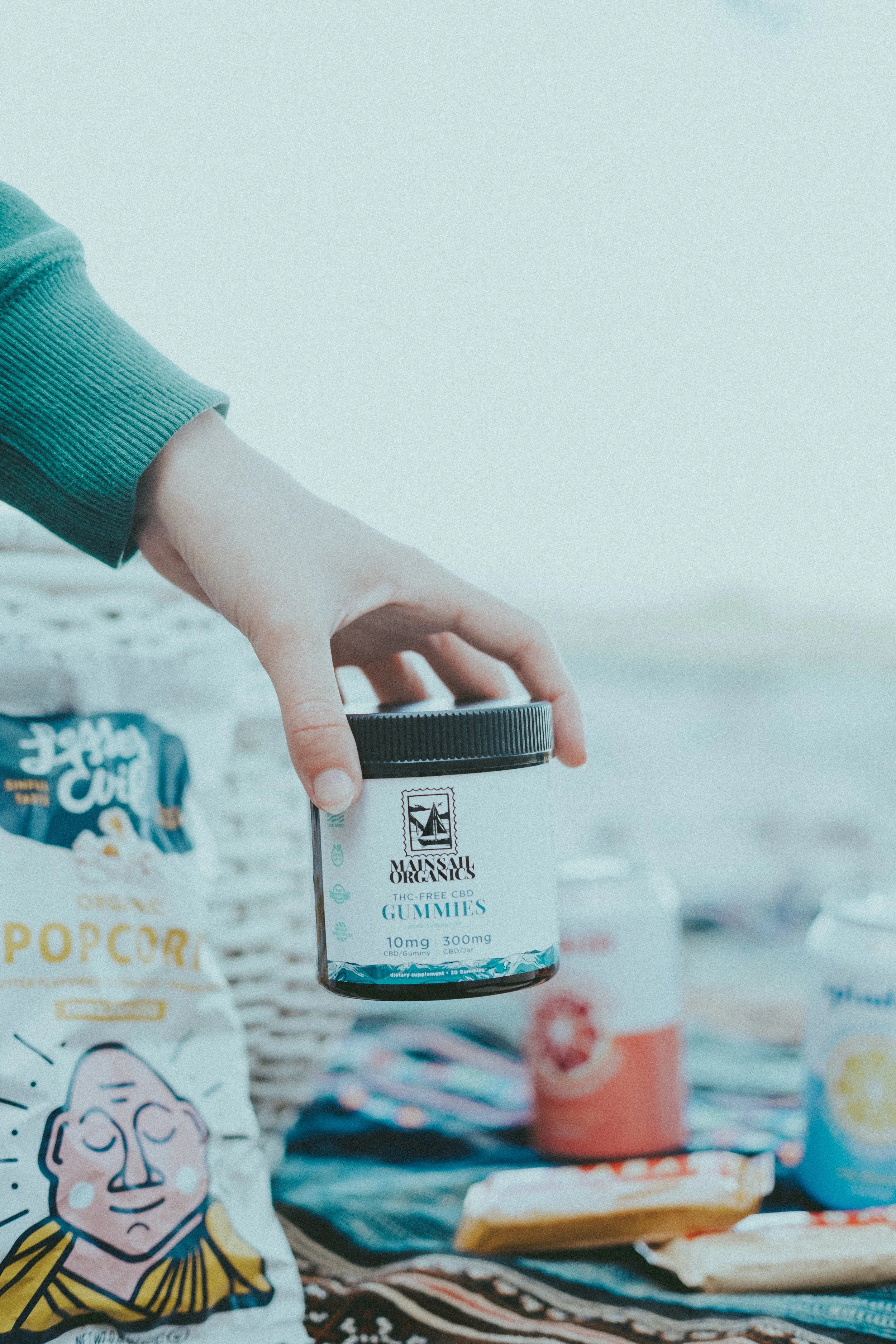 A hand holding a jar of MainSail Organics THC-free CBD gummies with various snack items on a blanket or surface nearby.