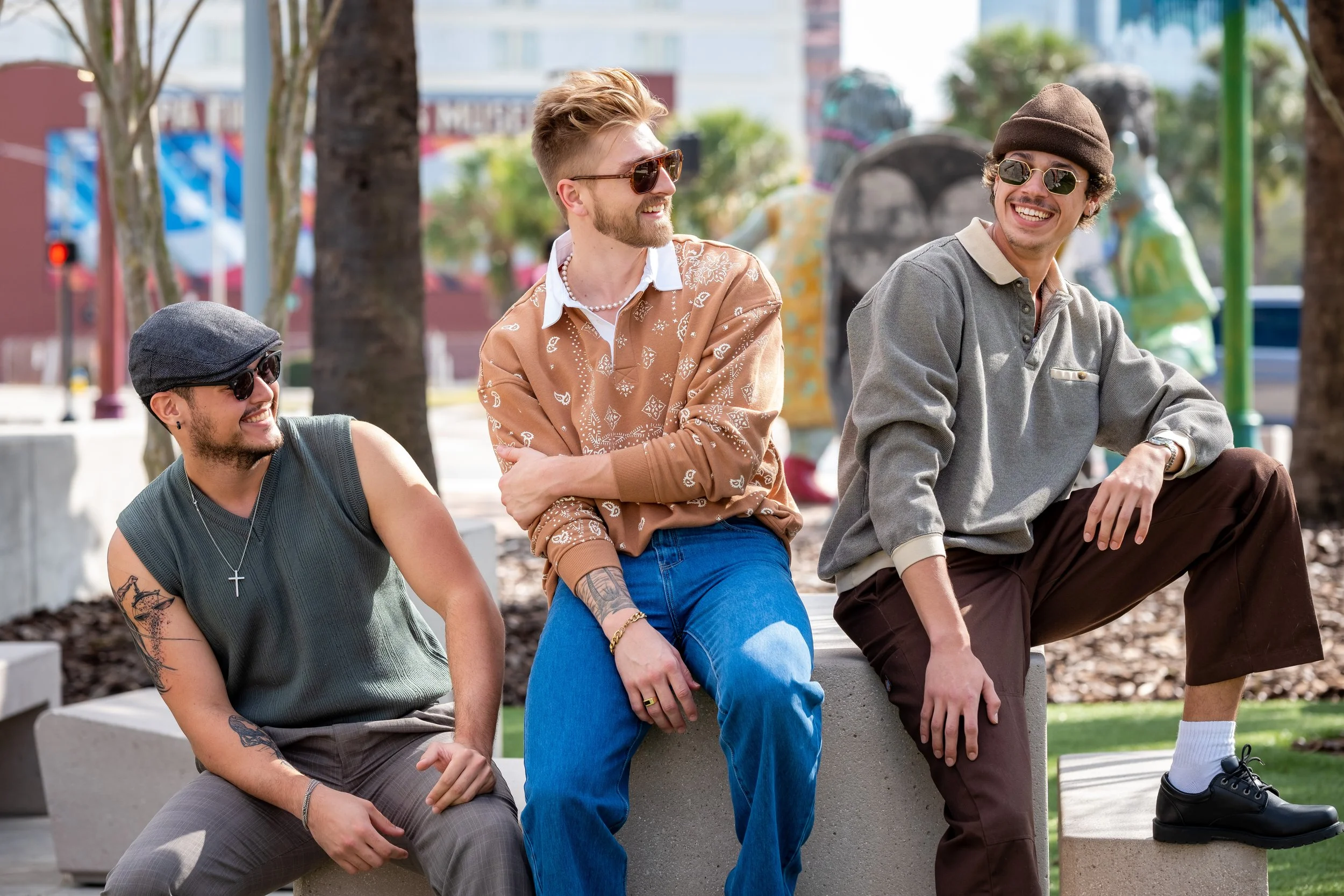 Three young men sitting outdoors on a sunny day, smiling and laughing, with trees and colorful sculptures in the background.