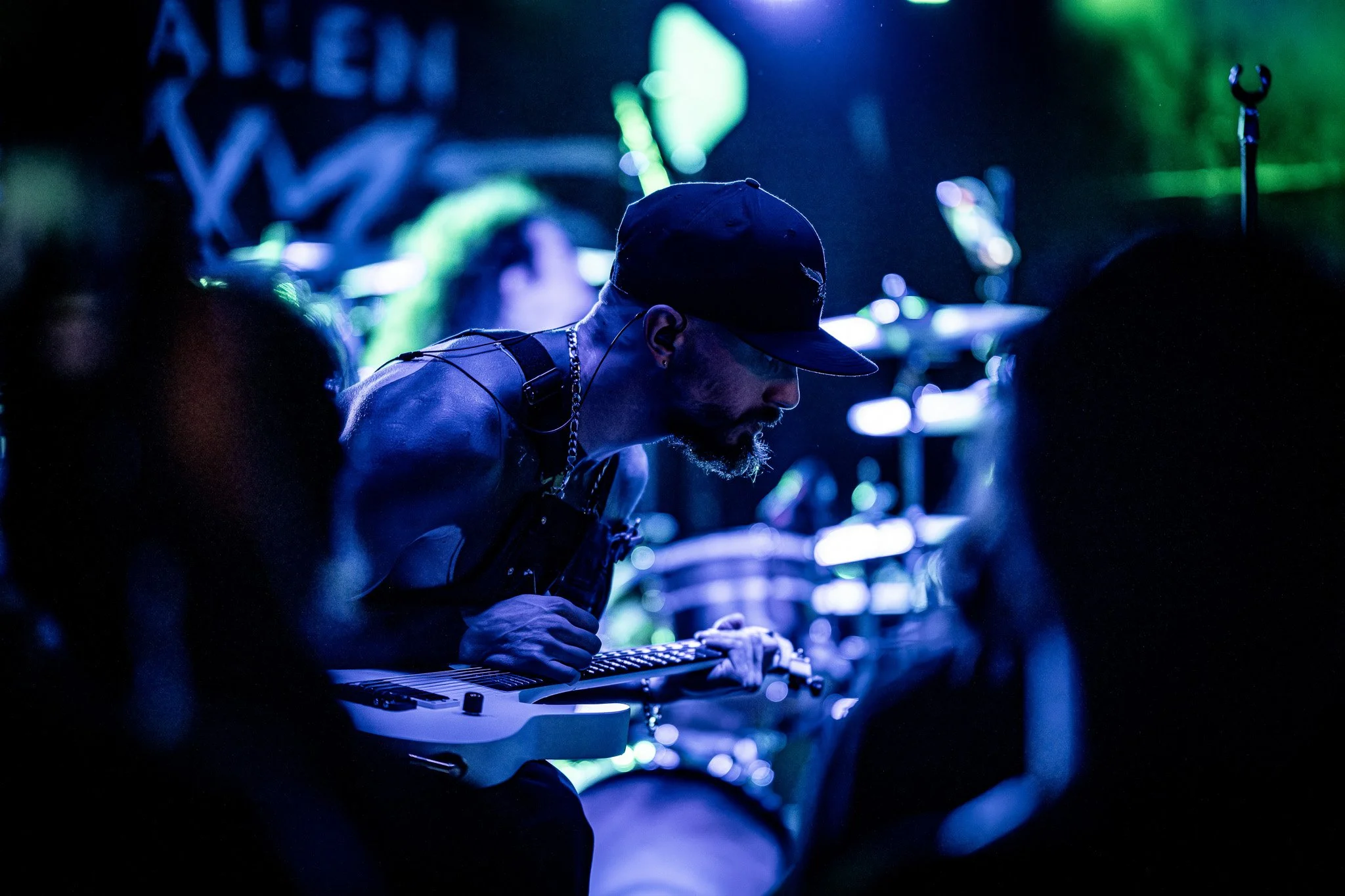 A musician wearing a black cap and sleeveless top playing an electric guitar on stage with blue and green lighting.