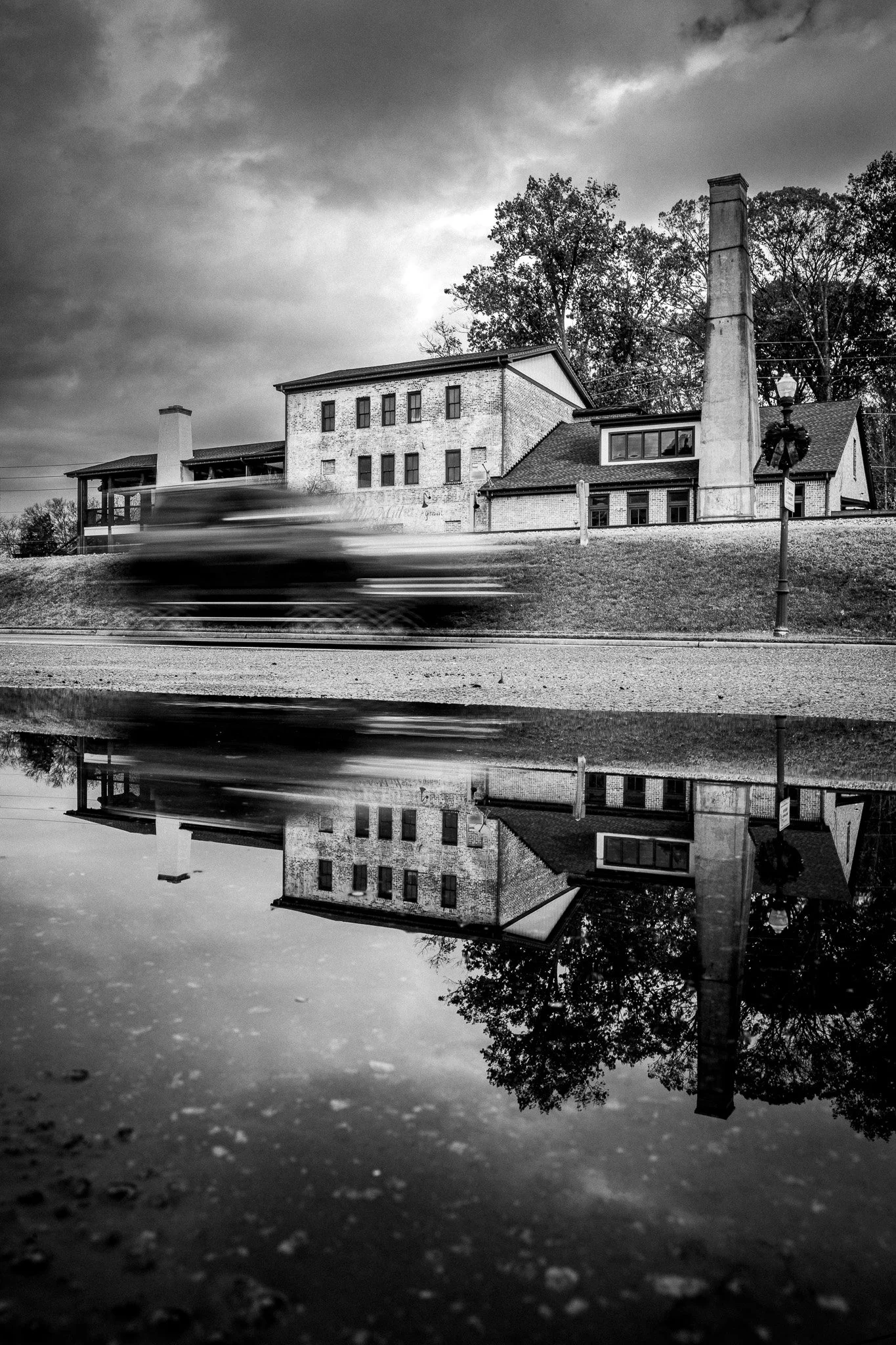 A black and white photo of an old brick house with a chimney and trees in the background. A blurred car is passing by, creating a reflection in a nearby puddle or water on the ground.