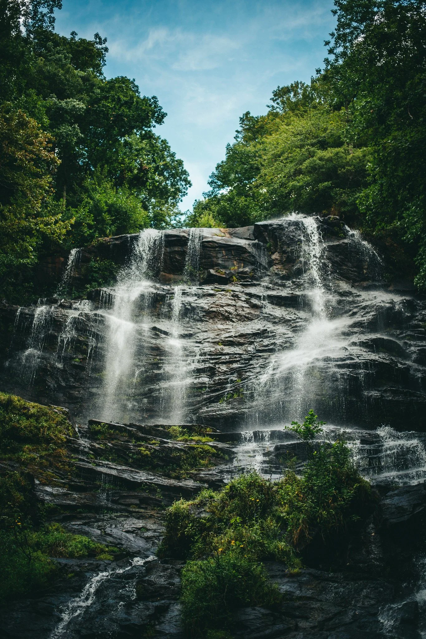 A waterfall flowing over rocks surrounded by green trees and a blue sky with some clouds.