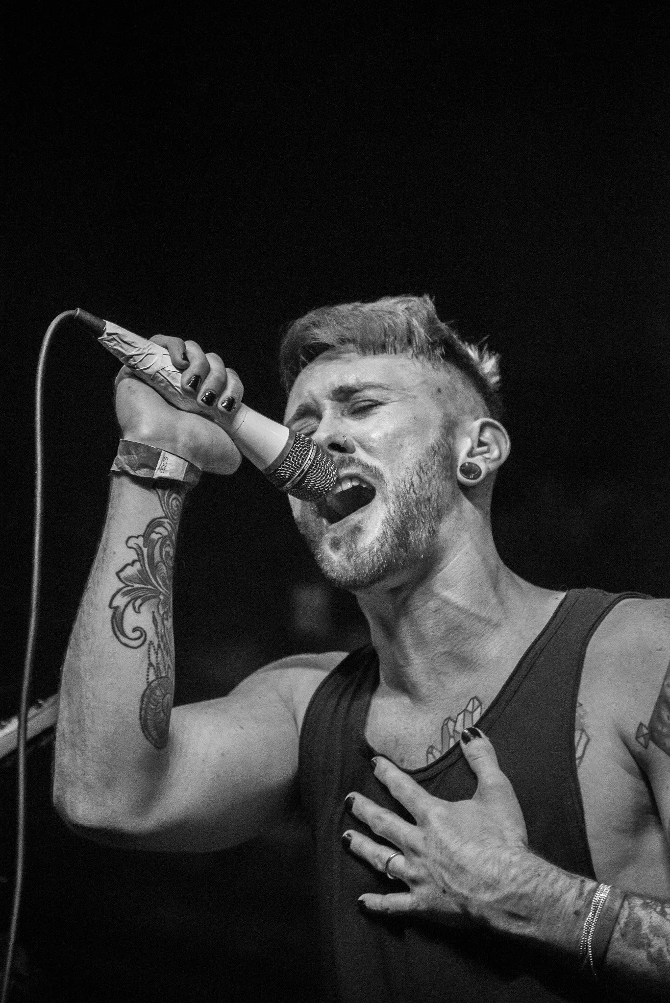 A black and white photo of a male singer passionately singing into a microphone, with one hand on his chest and the other holding the microphone, displaying tattoos on his arm and chest.