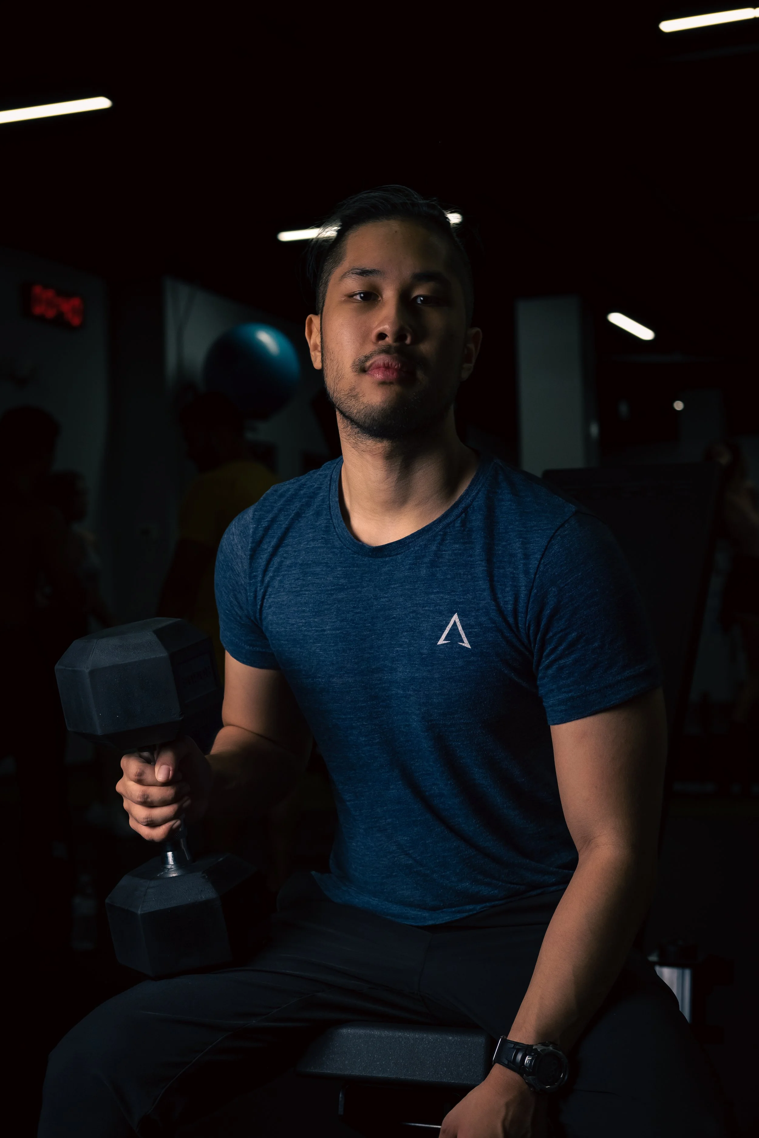 A man in a blue athletic shirt sitting in a gym, holding a dumbbell in his right hand, with gym equipment and other people working out in the background.