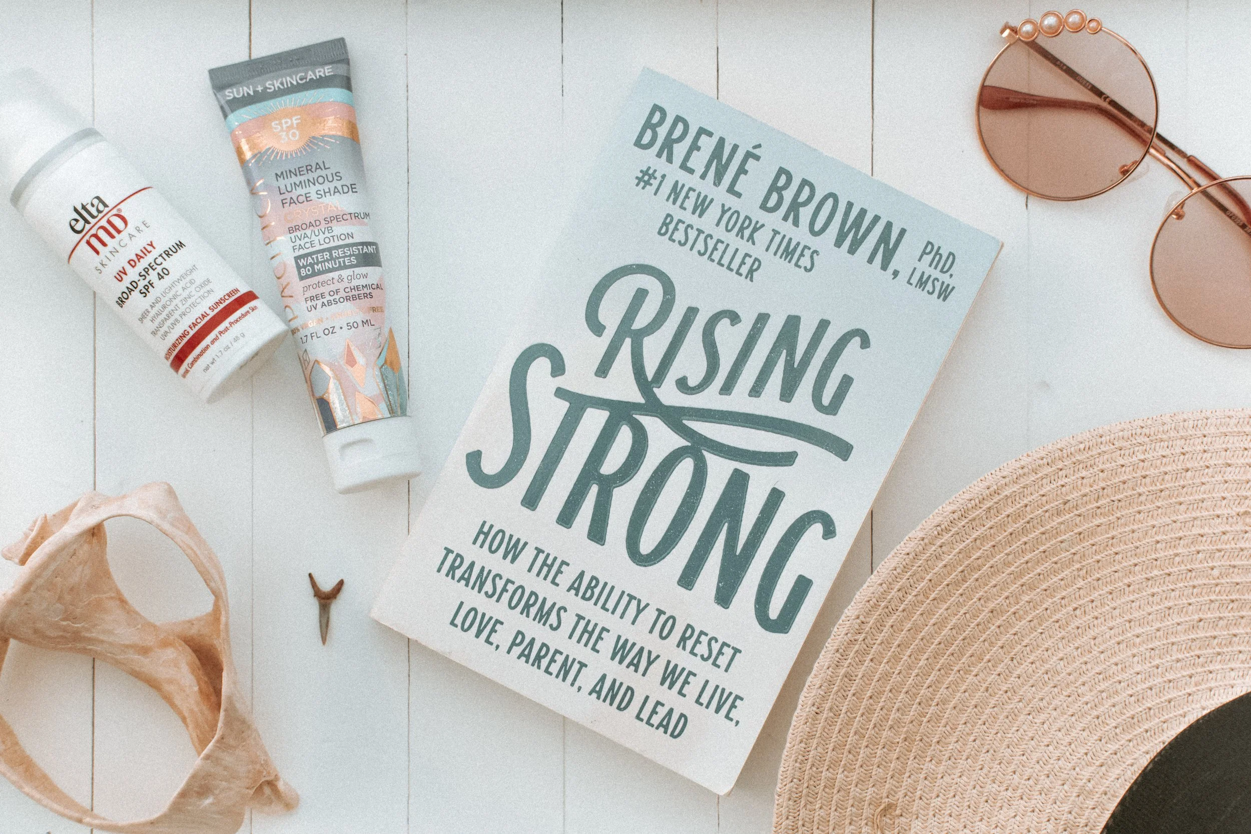 A book titled 'Rising Strong' by Brené Brown is surrounded by skincare products, a pair of round sunglasses, a woven hat, and a seashell on a white wooden surface.