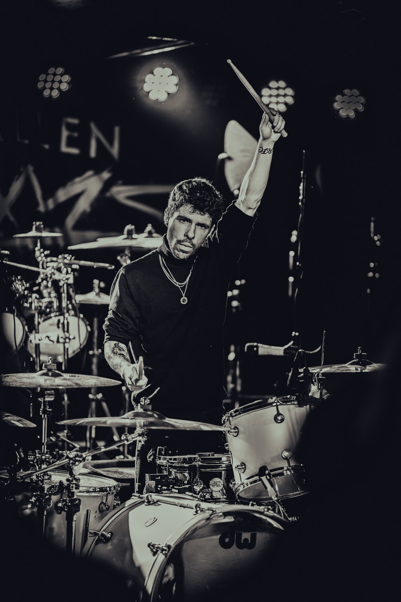 A male drummer playing a drum set on stage, illuminated by stage lights, in black and white.