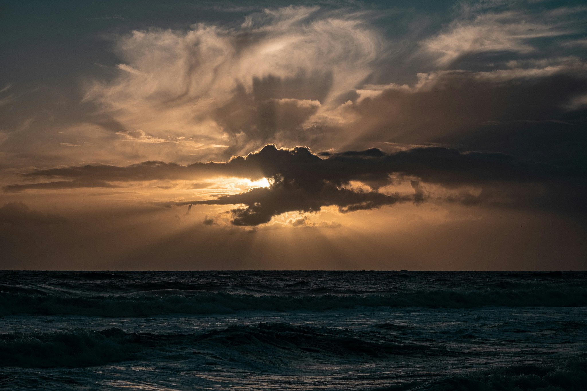 A sunset over the ocean with dark clouds and rays of sunlight breaking through