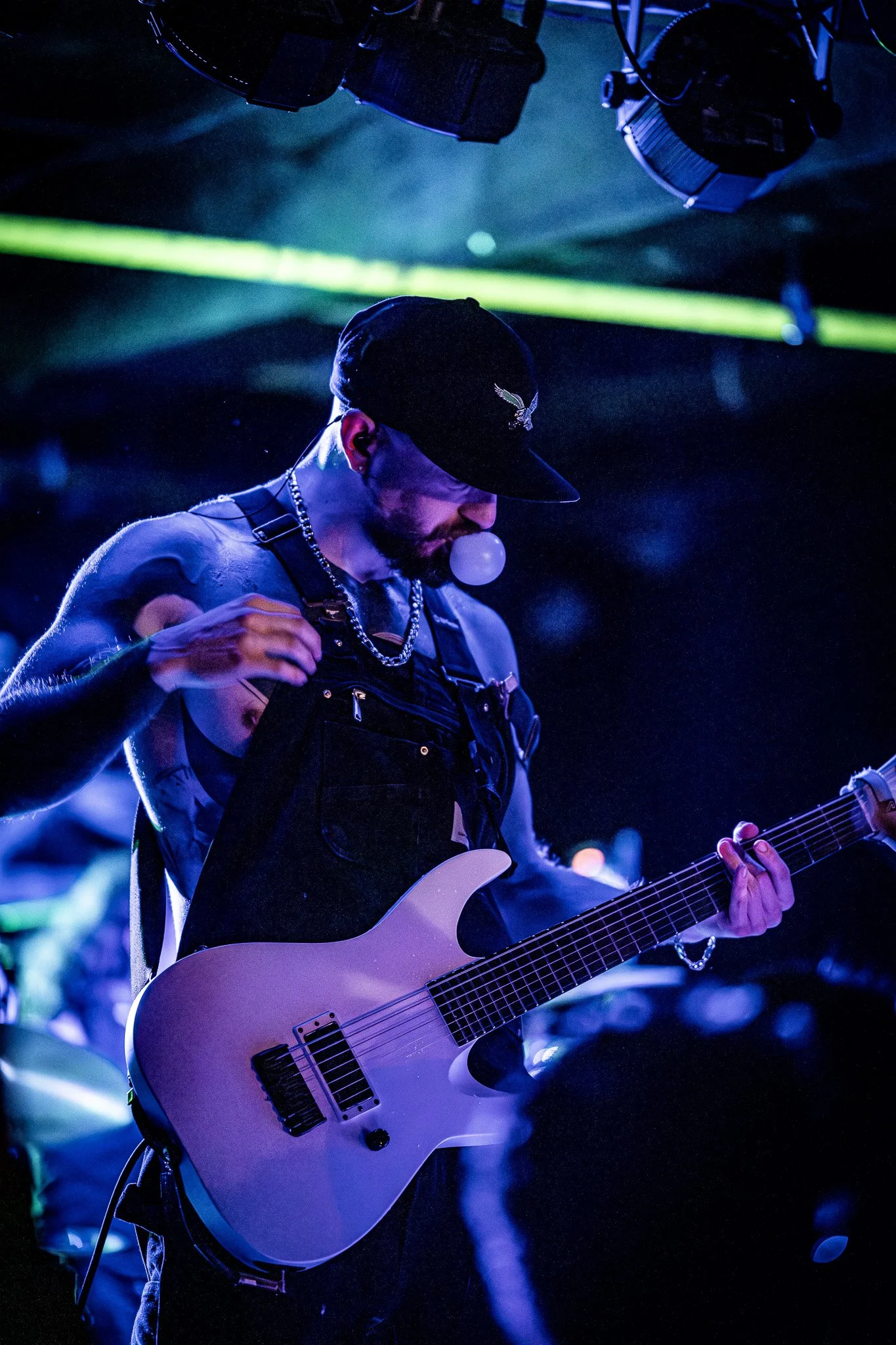 A male guitarist performing on stage with purple lighting and a dark background.