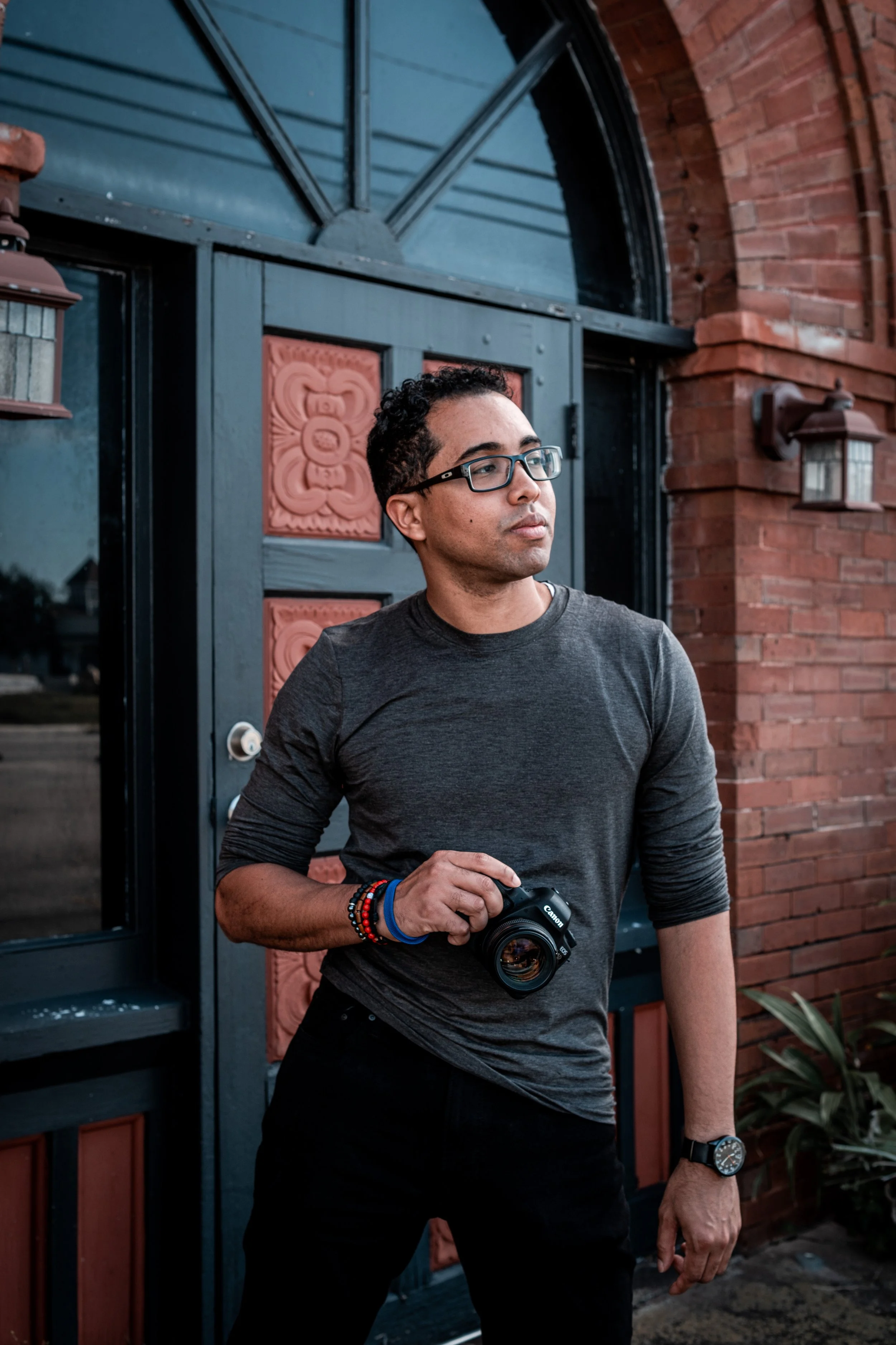A young man with dark curly hair and glasses stands outside a building holding a camera, dressed in a dark gray long-sleeve shirt and black pants, beside a brick wall with decorative stone panels.
