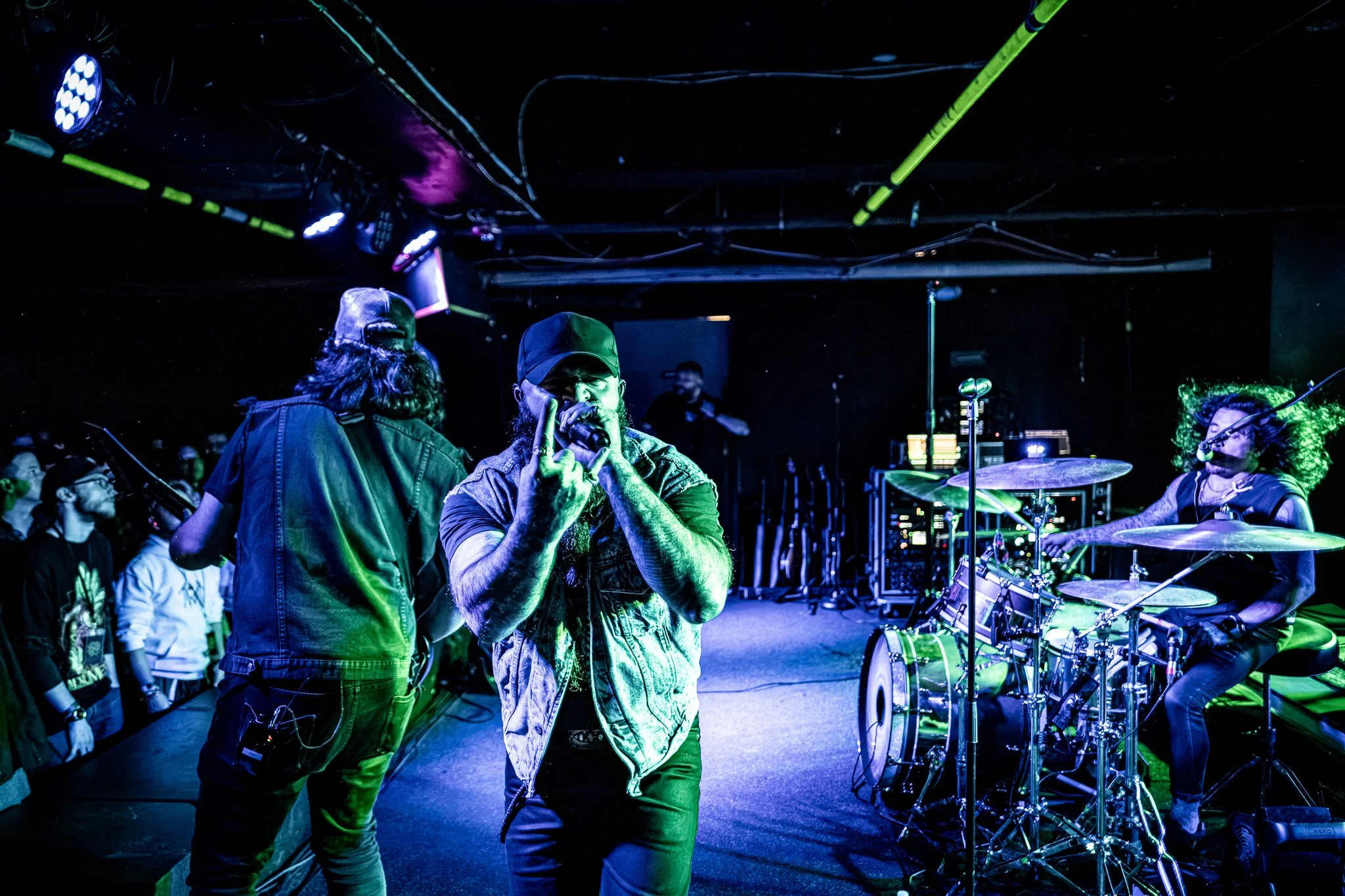 Live music performance on stage with male singer holding microphone, female drummer playing drums, and audience members watching, illuminated by colorful stage lights in a dark venue.