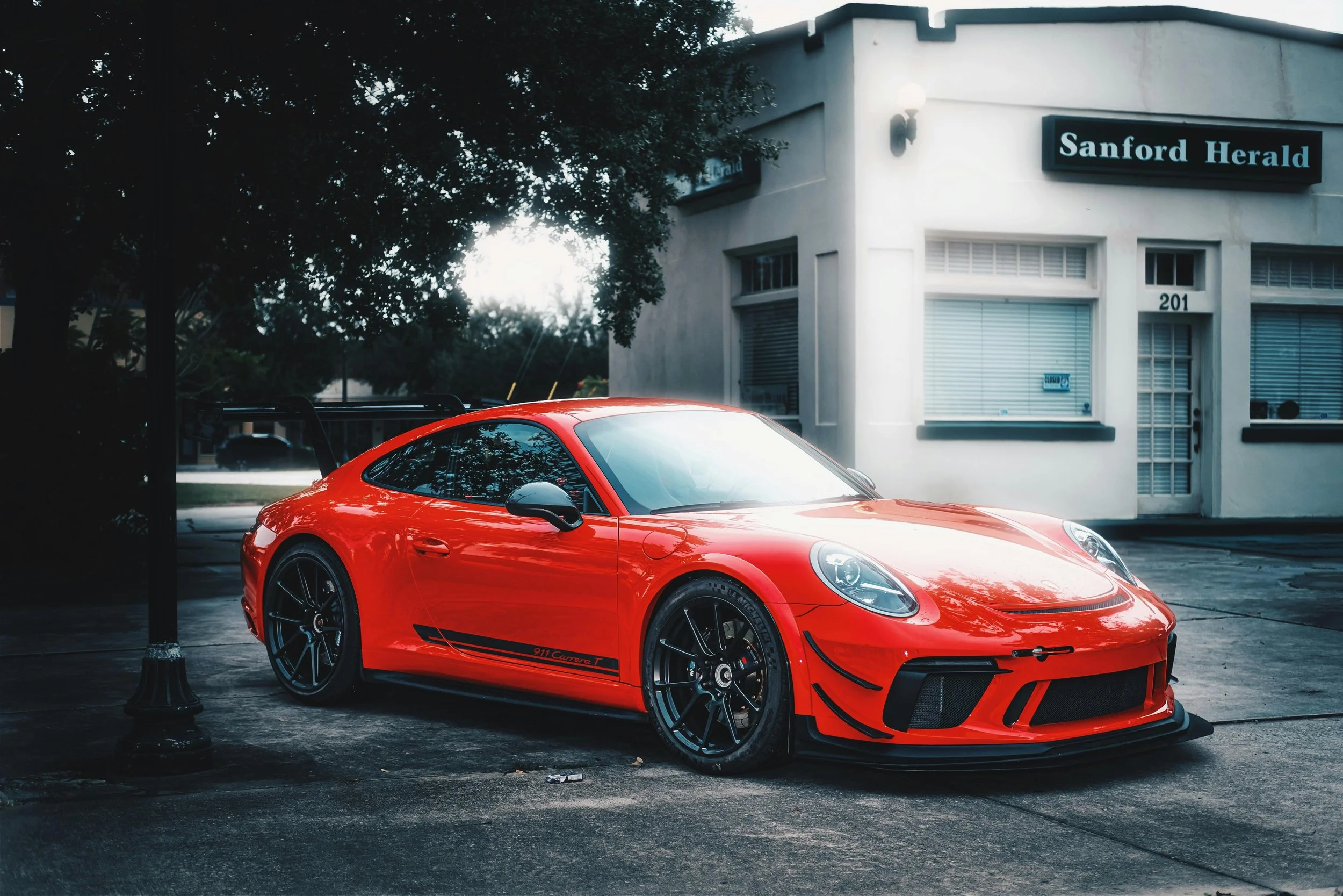 Red Porsche 911 Carrera T parked on the street near a tree and a white building with a sign that says 'Sanford Herald'.