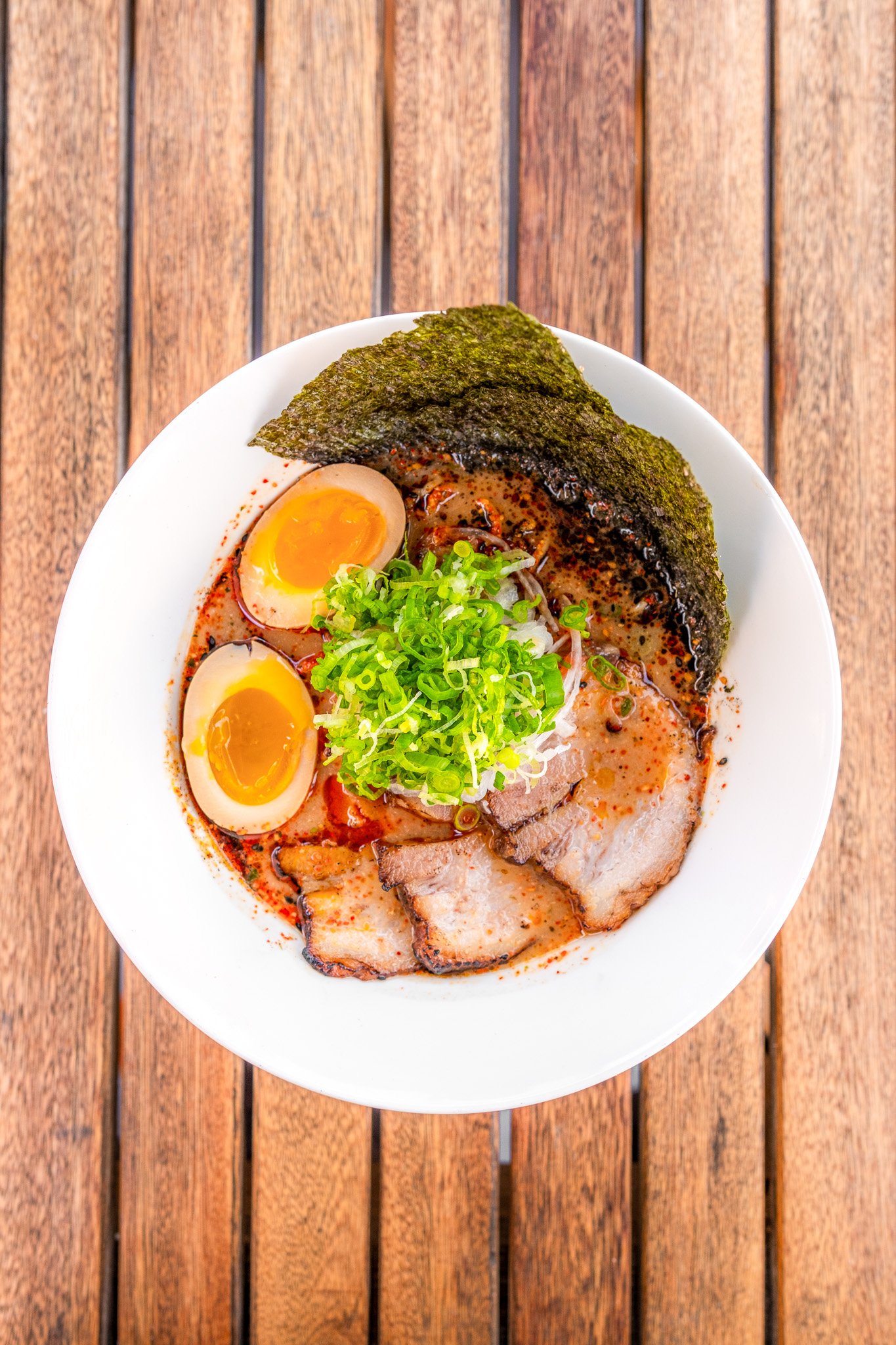 A bowl of ramen topped with soft-boiled eggs, sliced pork, chopped green onions, seaweed, and chili flakes, placed on a wooden surface.