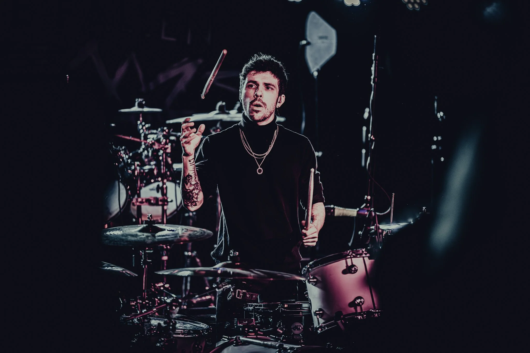A young male drummer performing on stage with a black shirt, layered necklaces, and tattoos on his arm, surrounded by a drum set and stage equipment.